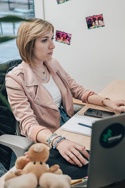 Une femme vêtue d'une veste rose est assise à un bureau, concentrée, avec un ordinateur portable, un bloc-notes et un smartphone. Des photos ornent le mur. Une peluche est au premier plan.