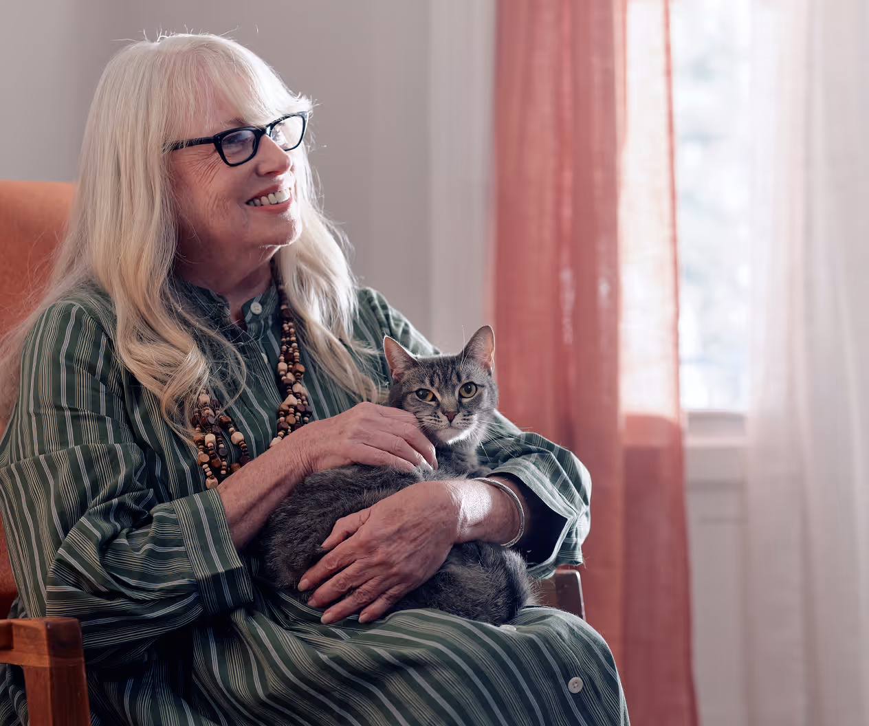 Smiling elderly woman with long white hair and glasses sitting in a chair holding a gray tabby cat.