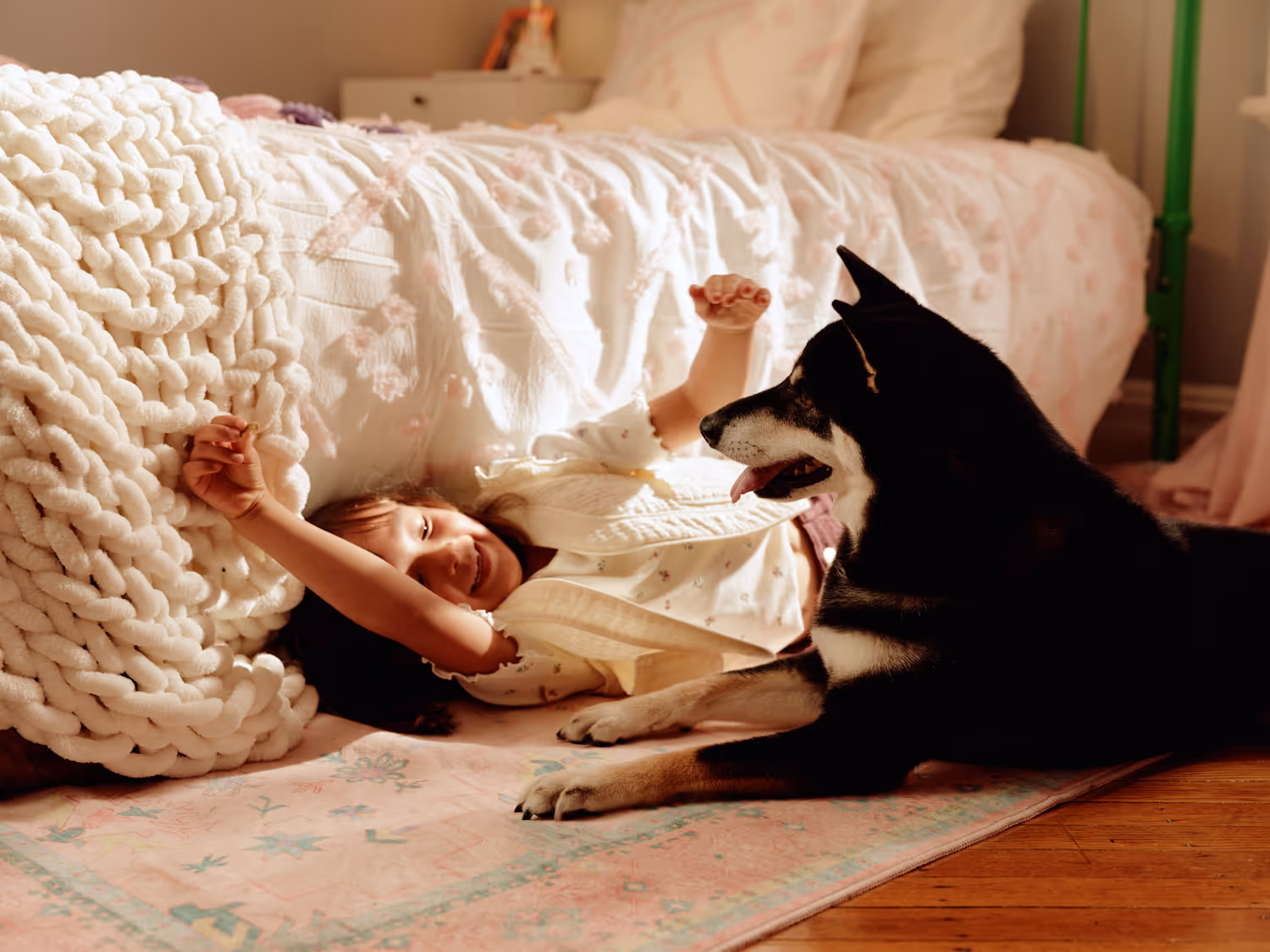 Young girl lying on a bedroom floor smiling and reaching out to a black and white dog lying nearby.