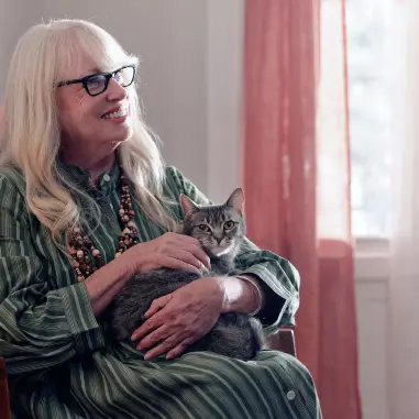 Smiling older woman with glasses holding a gray tabby cat on her lap near a window with sheer curtains.