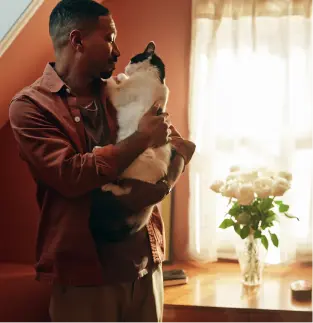 Man in brown shirt holding a black and white cat near a window with flowers on a table.