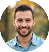 Smiling young man with short dark hair and beard wearing a light blue shirt against a blurred outdoor background.