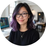 Smiling woman with shoulder-length black and purple hair wearing glasses and a black top in a well-lit office.