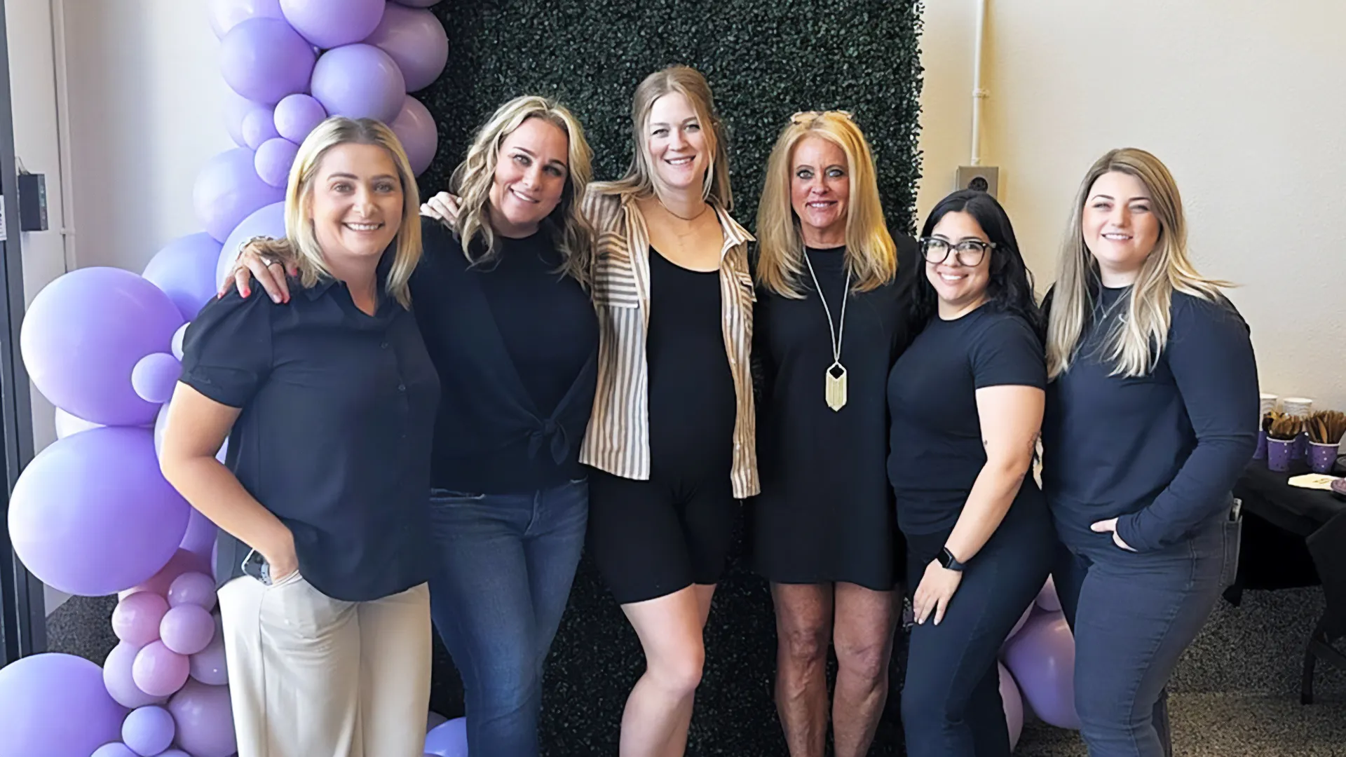 A group of six women smiling in front of a green foliage backdrop with purple balloon decorations. They are dressed casually, conveying a cheerful and celebratory atmosphere.