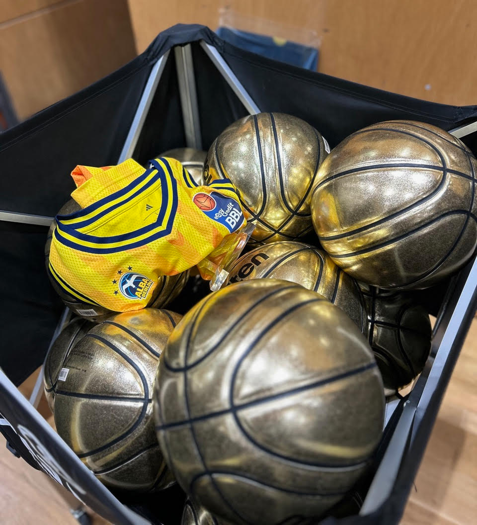 Korb mit mehreren goldfarbenen Basketbällen und einem gefalteten gelben Basketballtrikot mit dem Logo Alba Berlin.