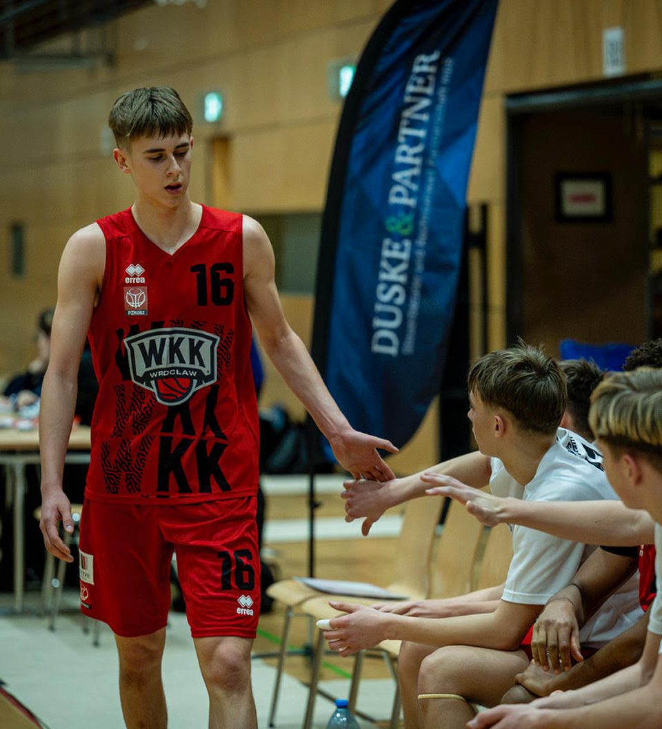 Basketballspieler in rotem Trikot mit der Nummer 16 reicht Teamkollegen und Mitspielern auf der Bank die Hand in einer Sporthalle.