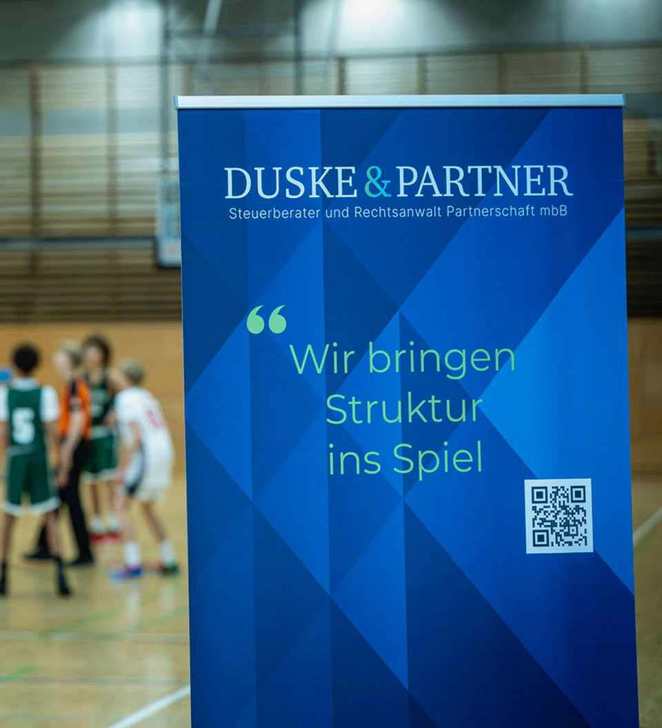 Blauer Werbebanner in einer Sporthalle mit dem Text „DUSKE & PARTNER Steuerberater und Rechtsanwalt Partnerschaft mbB“ und dem Slogan „Wir bringen Struktur ins Spiel“ sowie einem QR-Code, unscharfe Basketballspieler im Hintergrund.