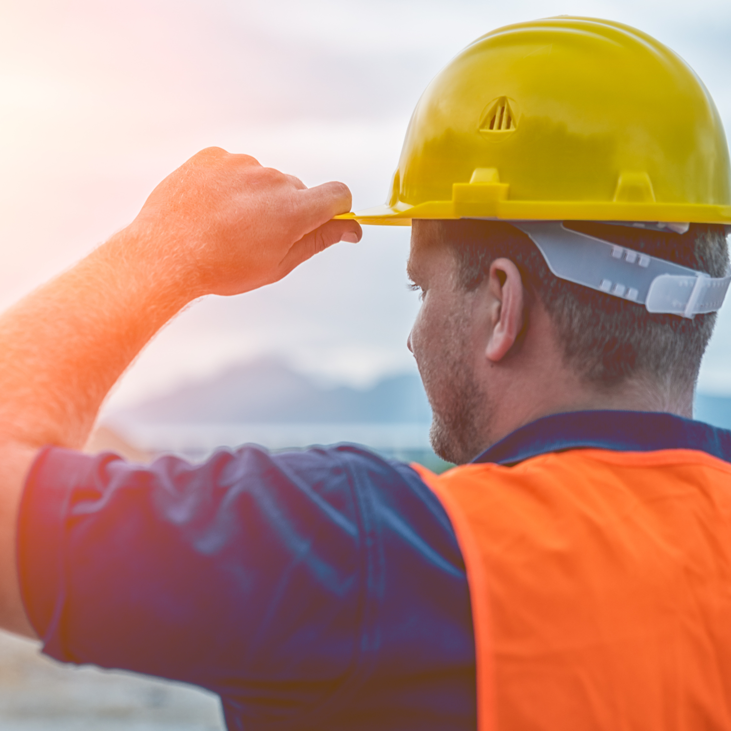 Construction worker wearing a yellow hard hat and orange safety vest adjusting his helmet outdoors at sunrise or sunset.