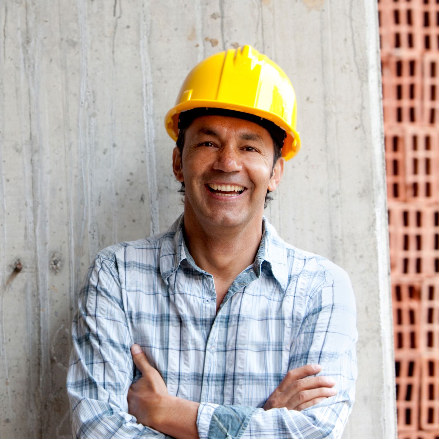 Smiling construction worker wearing a yellow hard hat and plaid shirt, standing with arms crossed against a concrete wall.