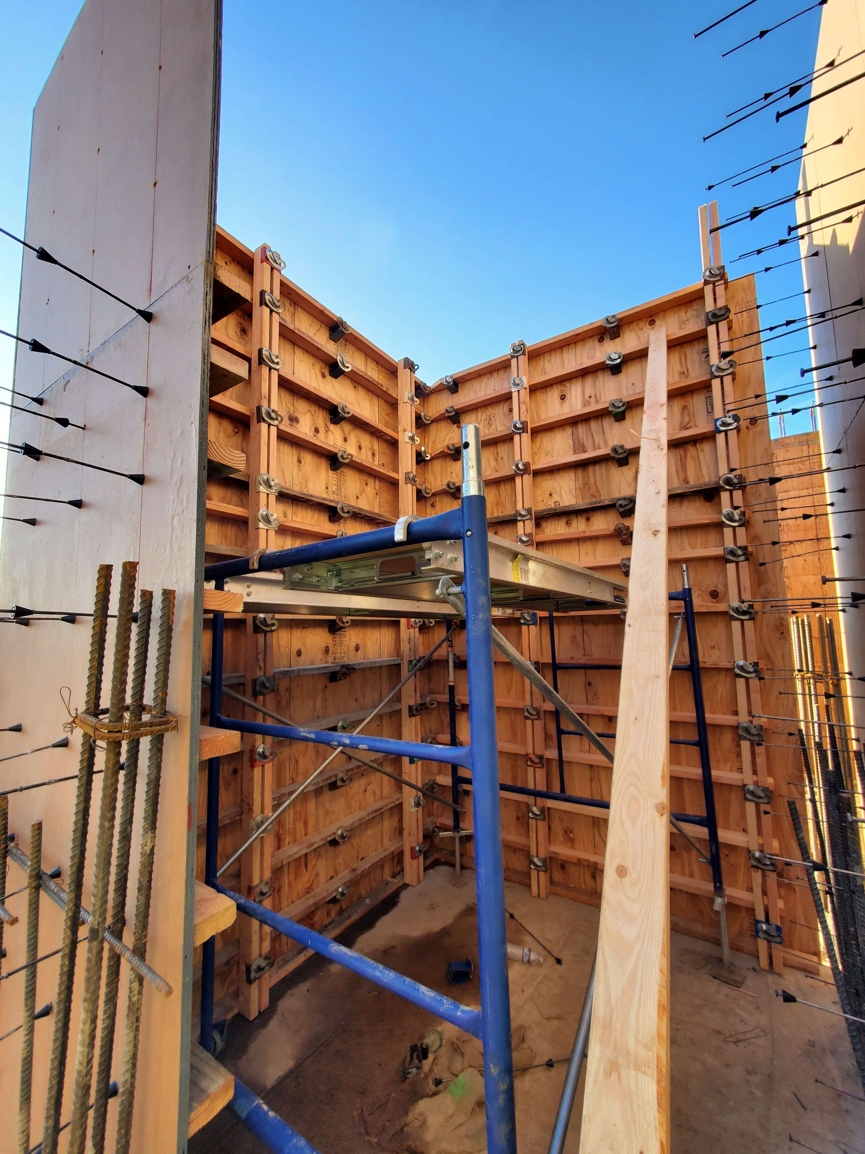 Construction site with wooden formwork, metal ties, and blue scaffolding under clear blue sky.