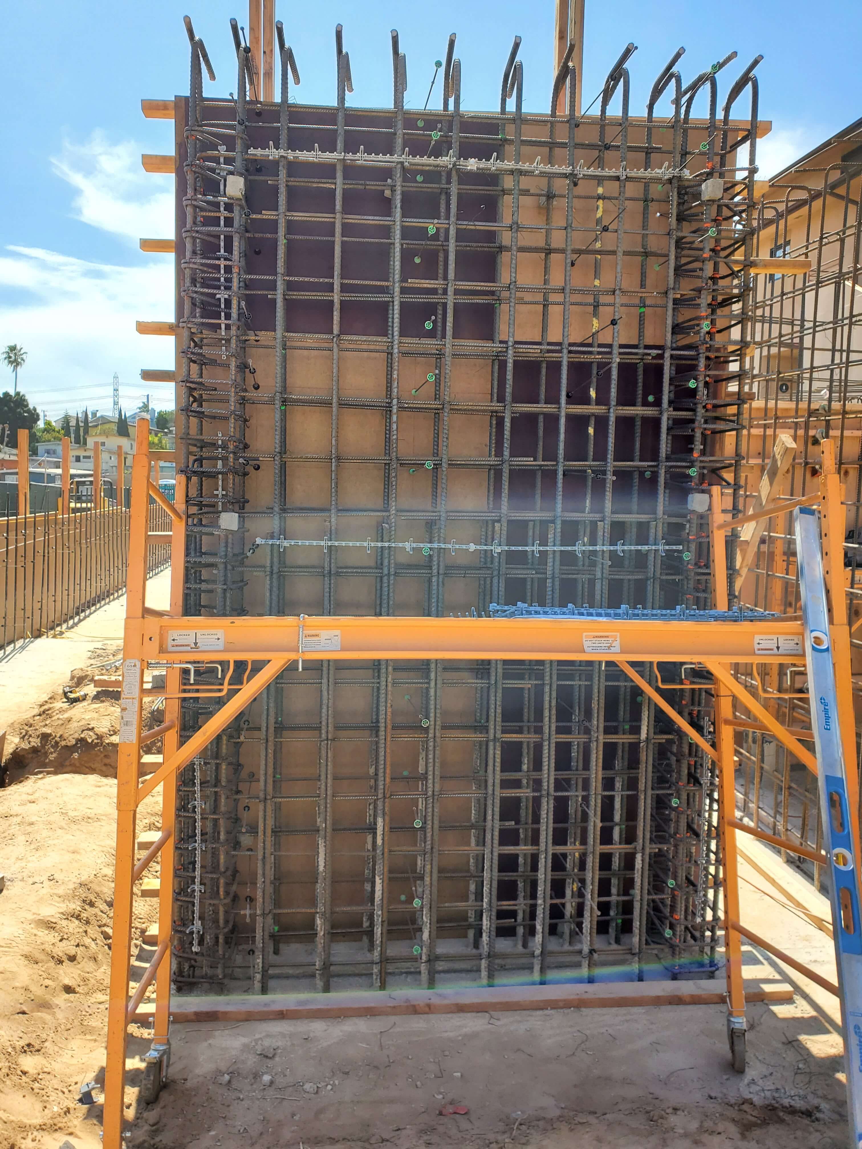 Steel rebar framework for a vertical concrete wall under construction with orange scaffolding and a ladder on a sunny construction site.