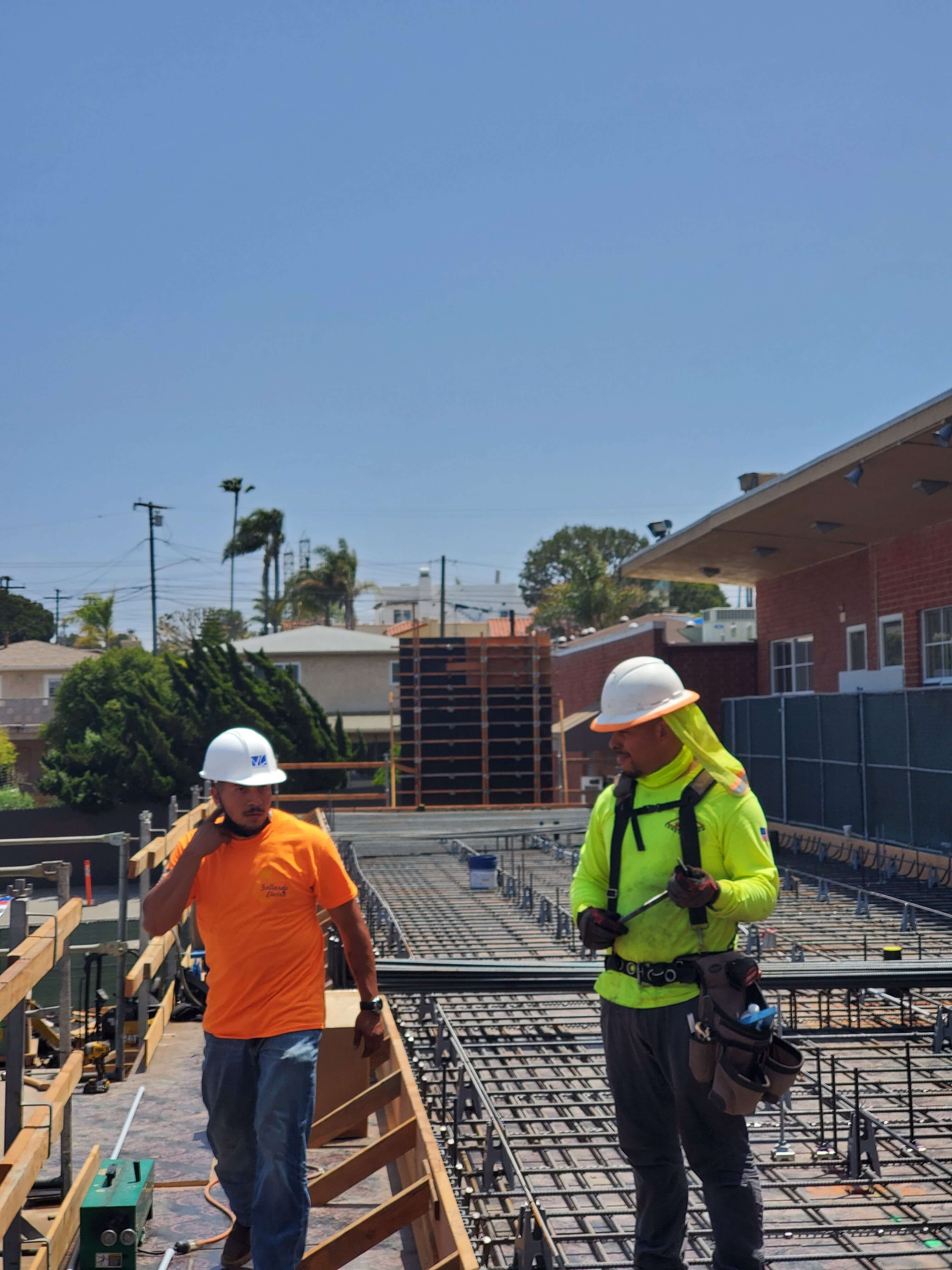 Two construction workers wearing safety helmets and bright shirts working on a steel rebar structure at a construction site.