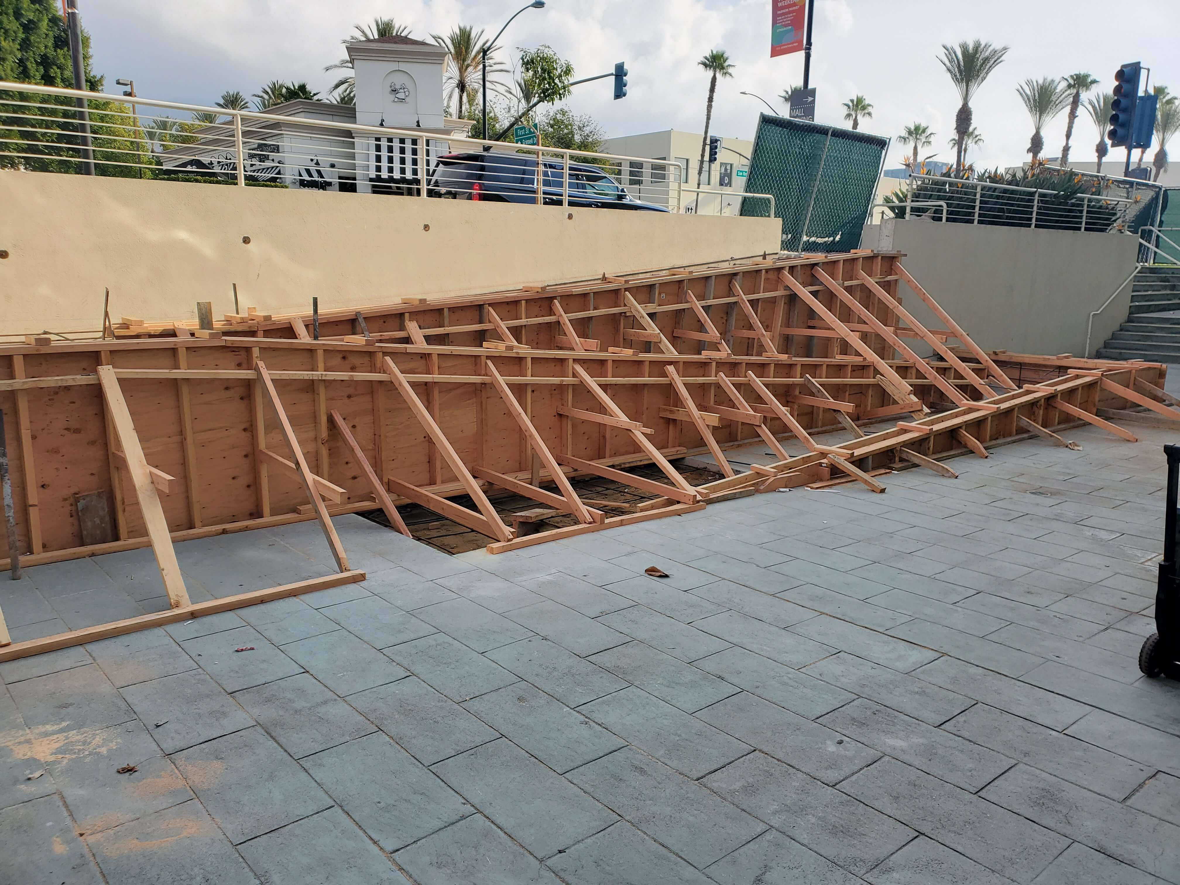 Wooden framework supporting a steep slope beside paved walkway and stairs with palm trees and buildings in the background.