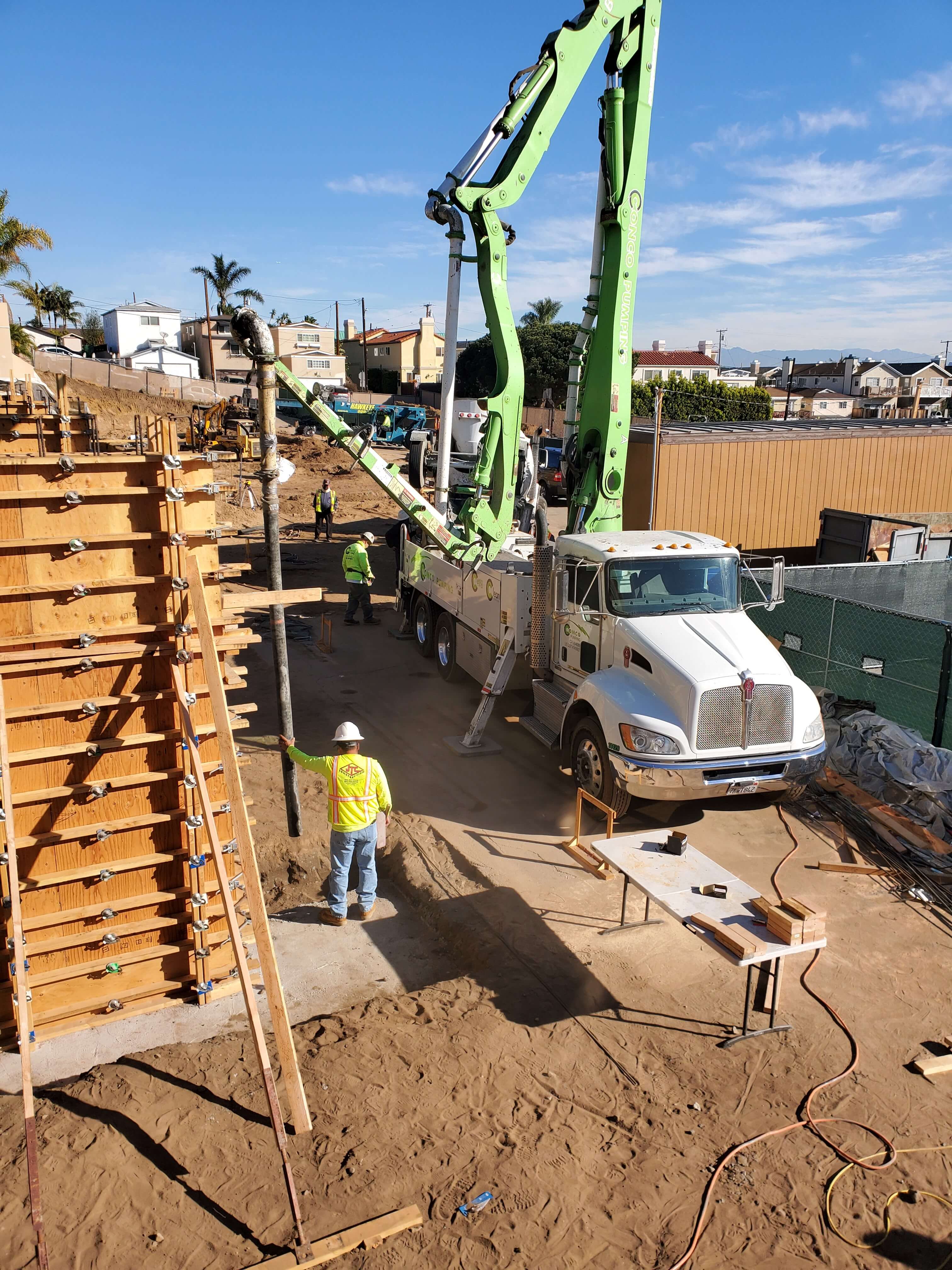 Construction site with workers operating a white concrete pump truck with green arms pouring concrete into wooden forms.