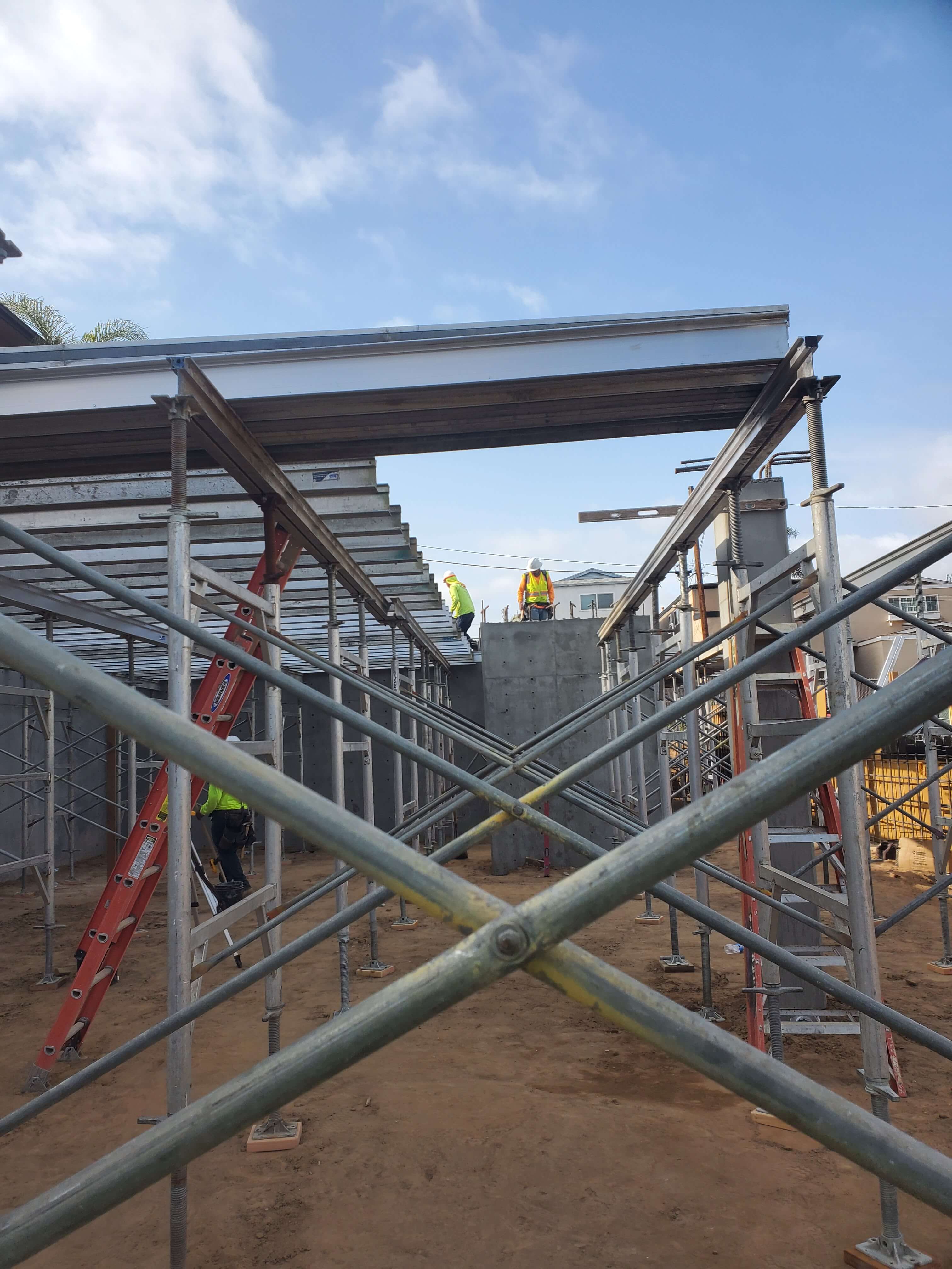 Construction site with scaffolding, steel braces, and workers in safety vests and helmets assembling a structure under a blue sky.