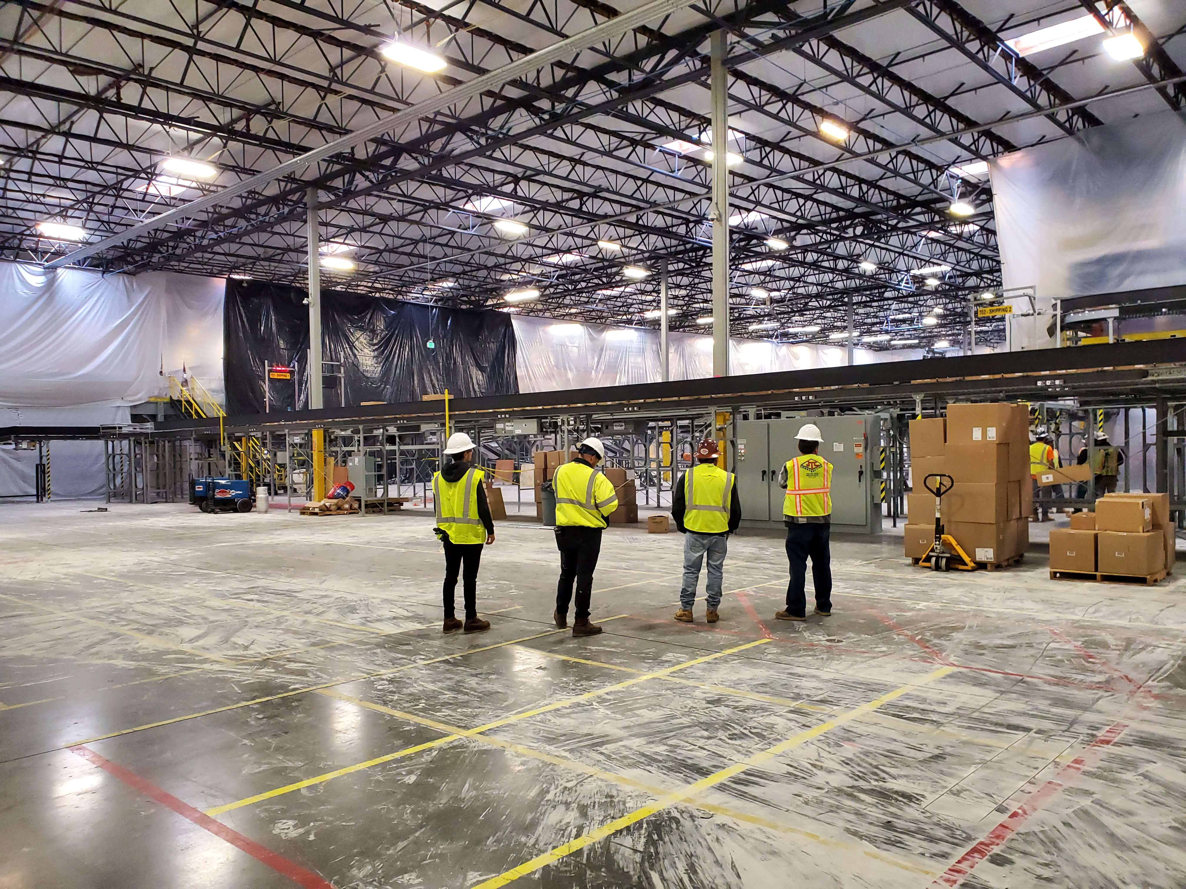 Four construction workers in safety vests and helmets standing inside a large, empty warehouse with industrial equipment and stacked cardboard boxes.