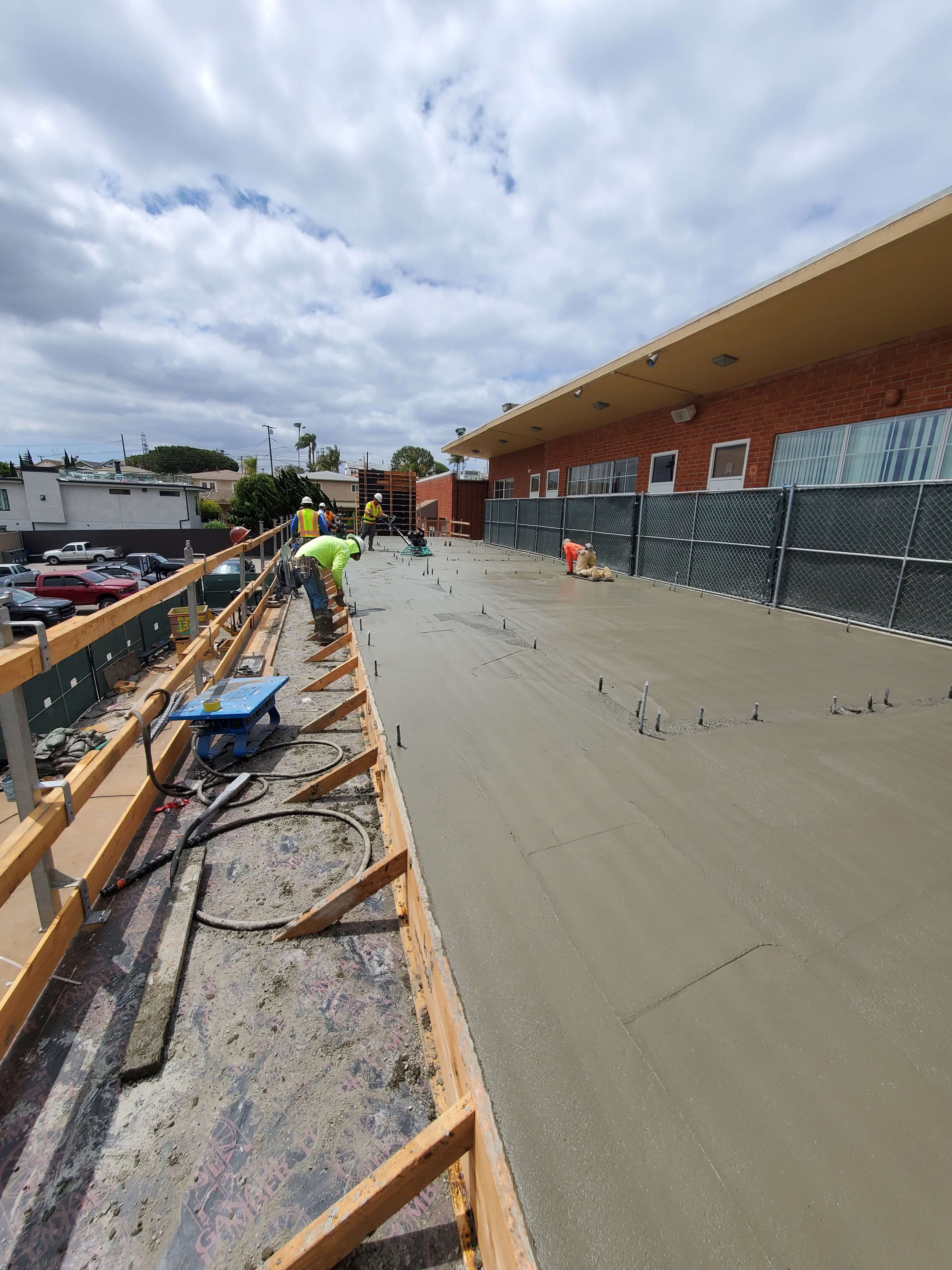 Construction workers wearing safety gear smoothing freshly poured concrete on a rooftop under a cloudy sky.