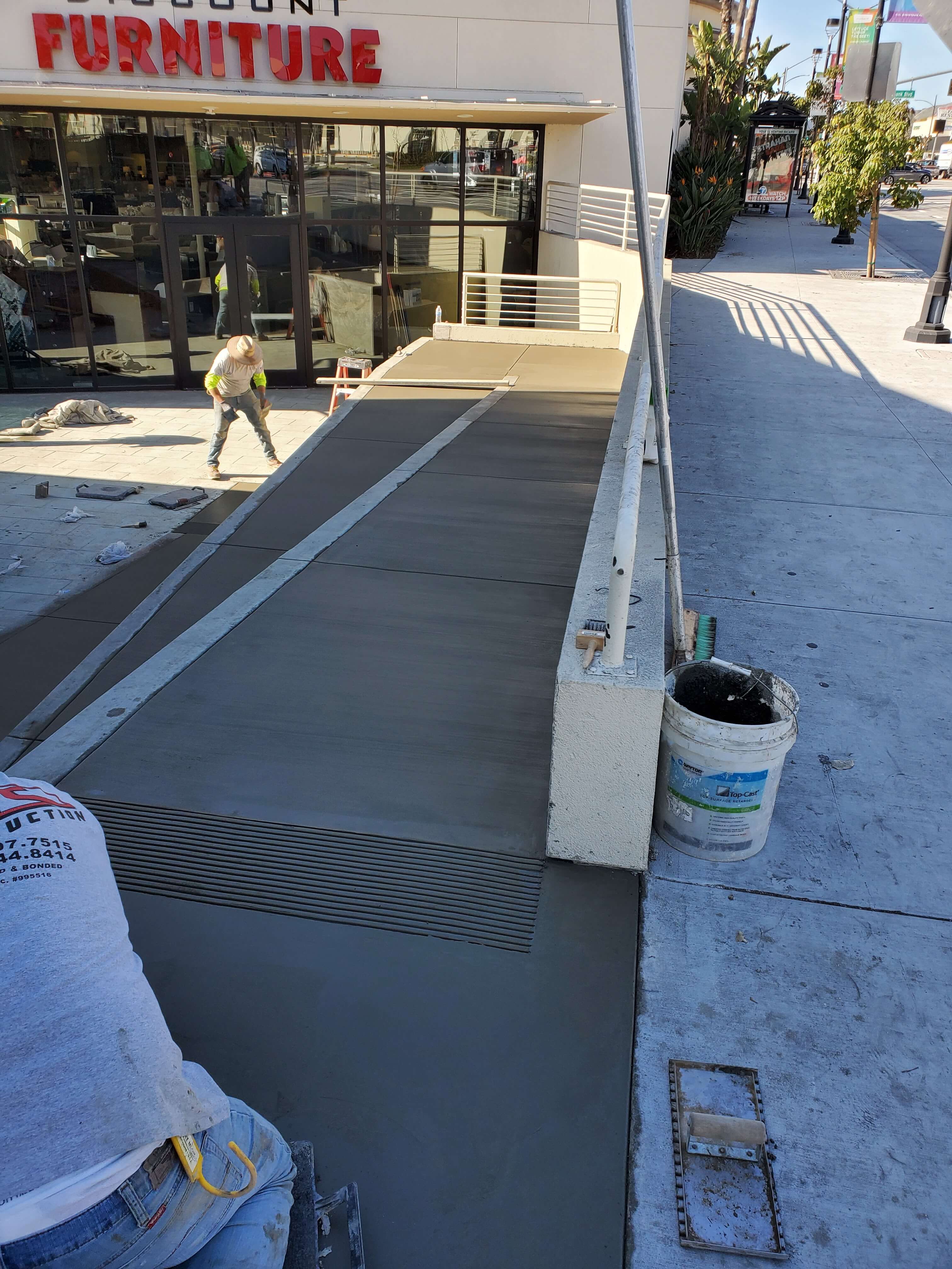 Workers smoothing freshly poured concrete on a ramp outside a furniture store under bright sunlight.