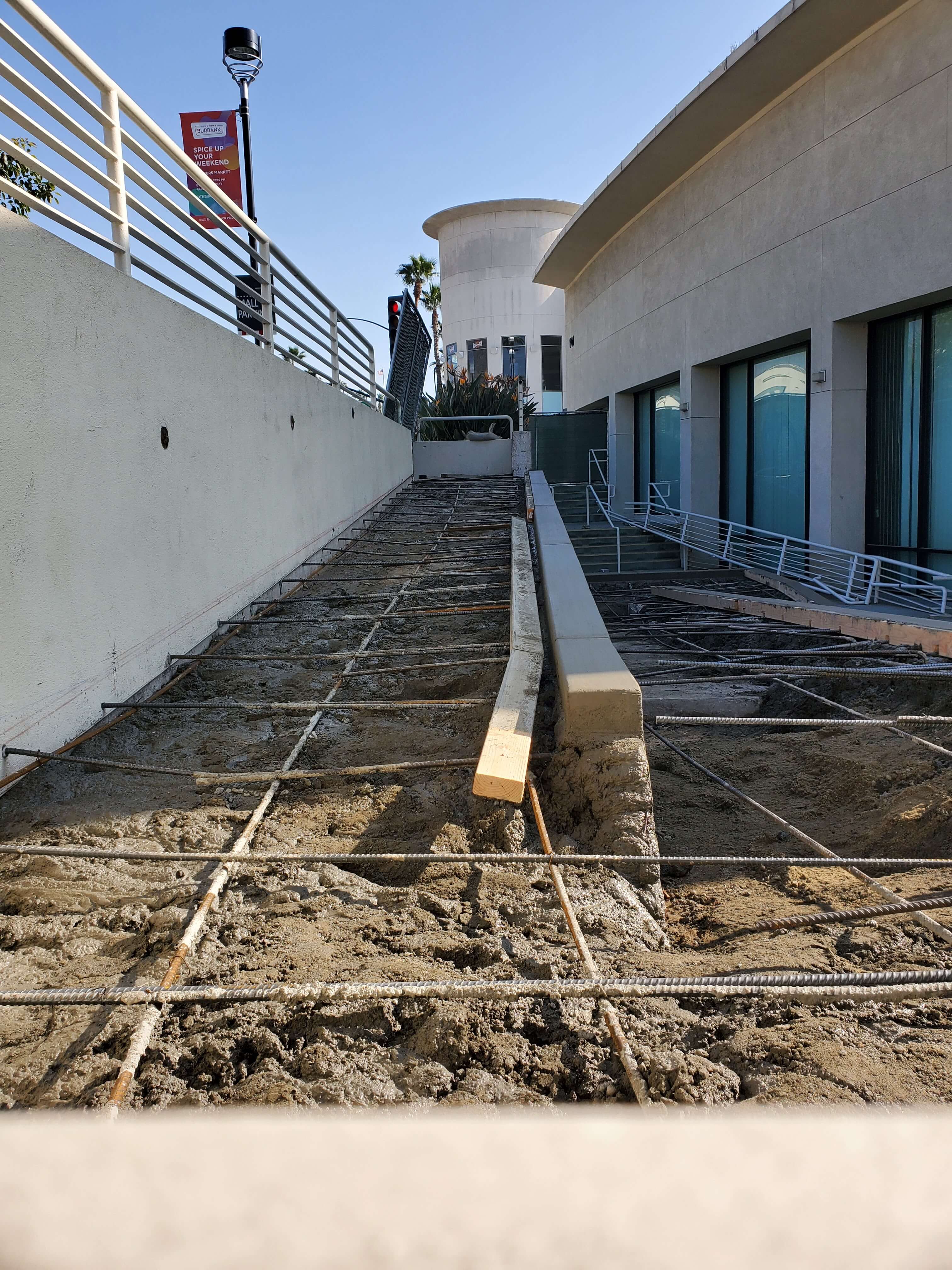 Concrete ramp under construction with metal rebar grid and wooden formwork between two buildings under clear blue sky.
