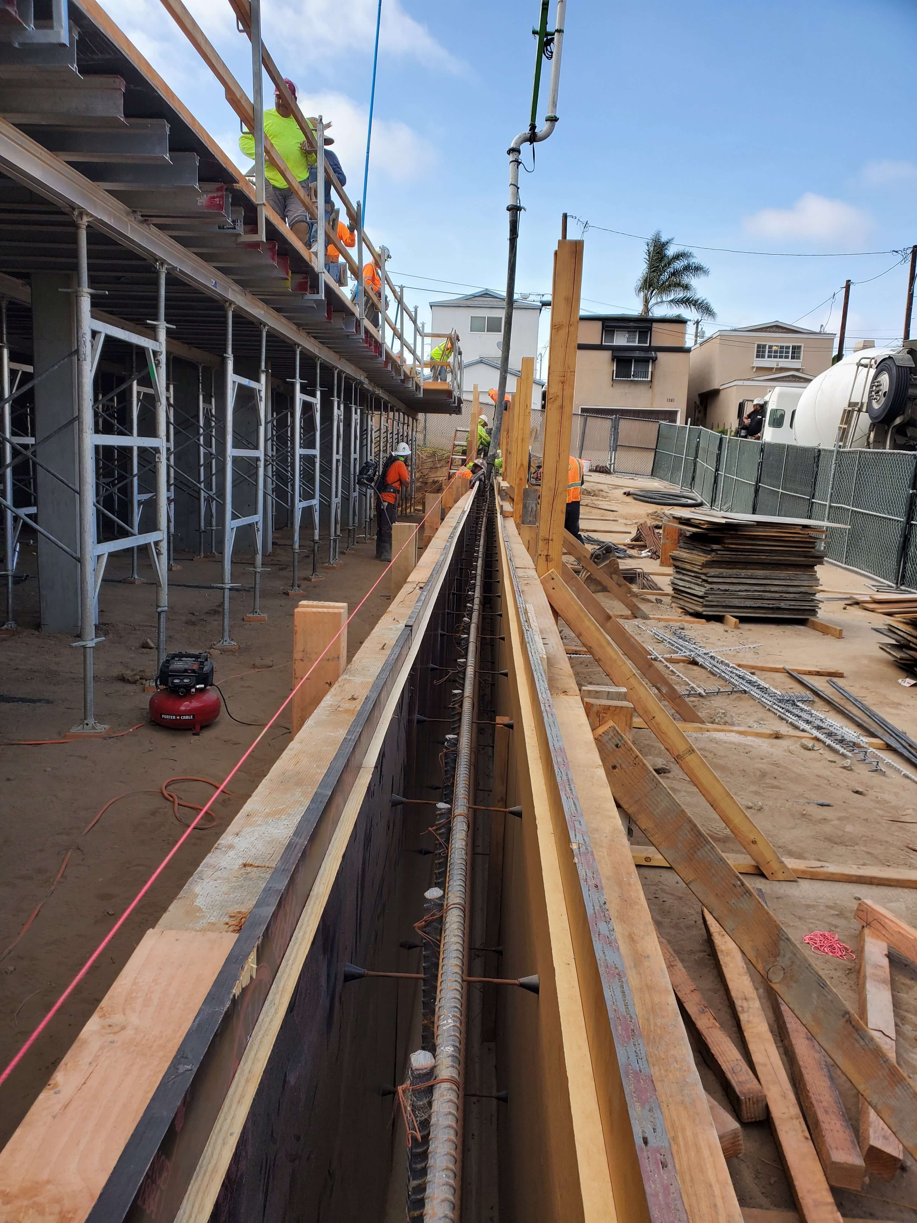 Construction site showing workers in safety gear building a long reinforced concrete wall form next to scaffolding.