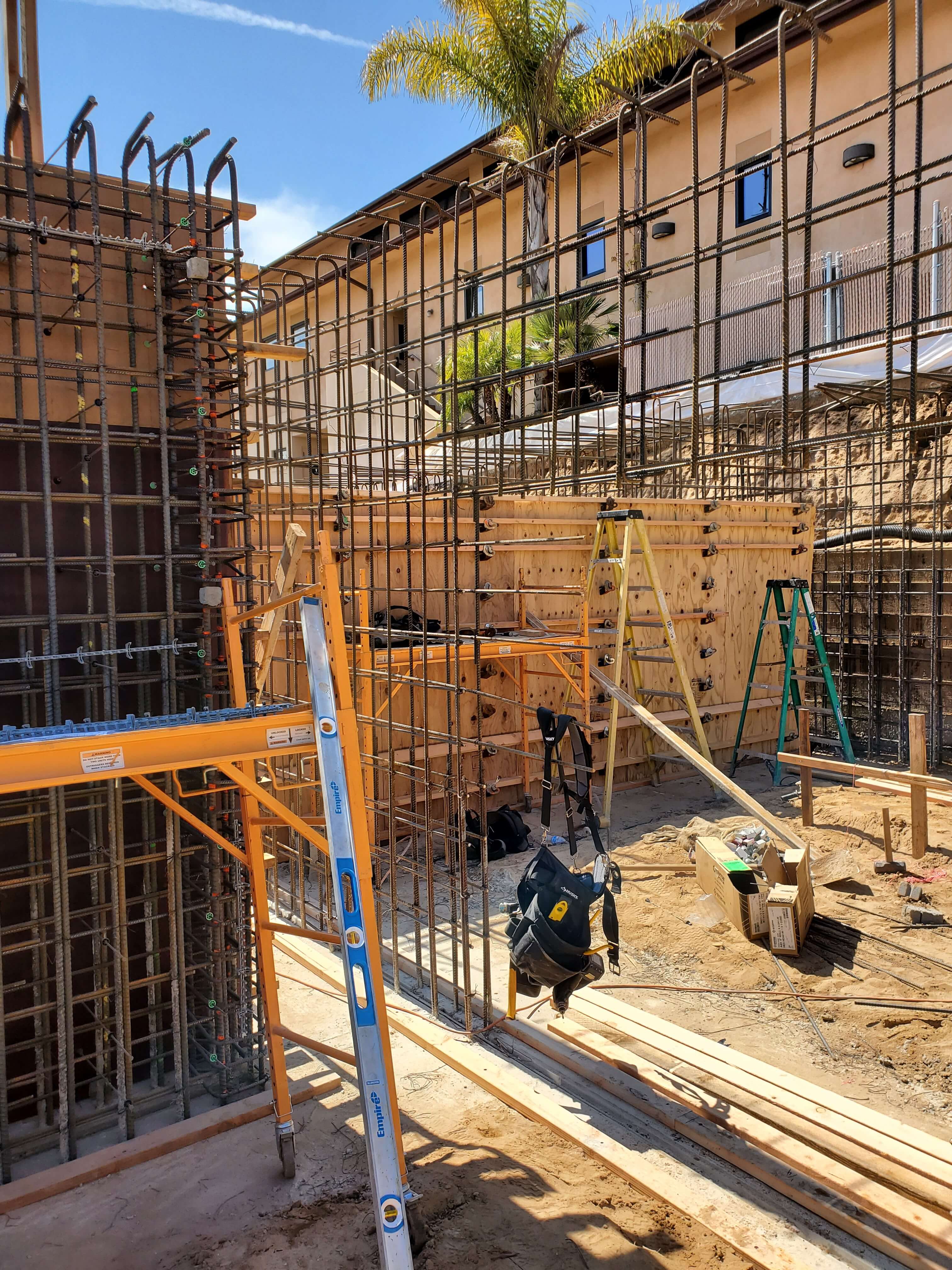 Construction site with metal rebar framework, wooden formwork, ladders, and scattered building materials under a clear blue sky.