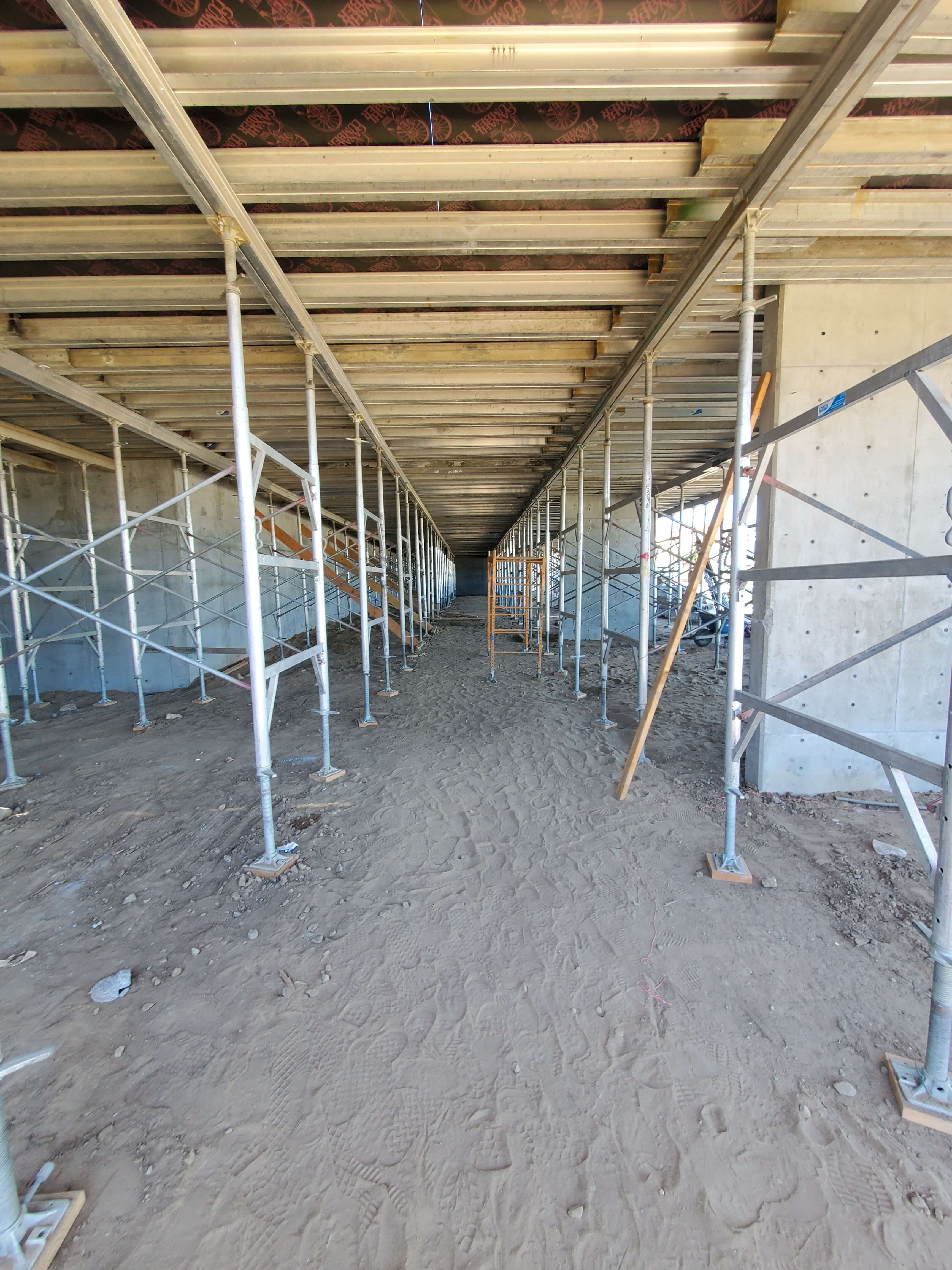 Construction site corridor with metal scaffolding supporting wooden beams and dirt floor with footprints.