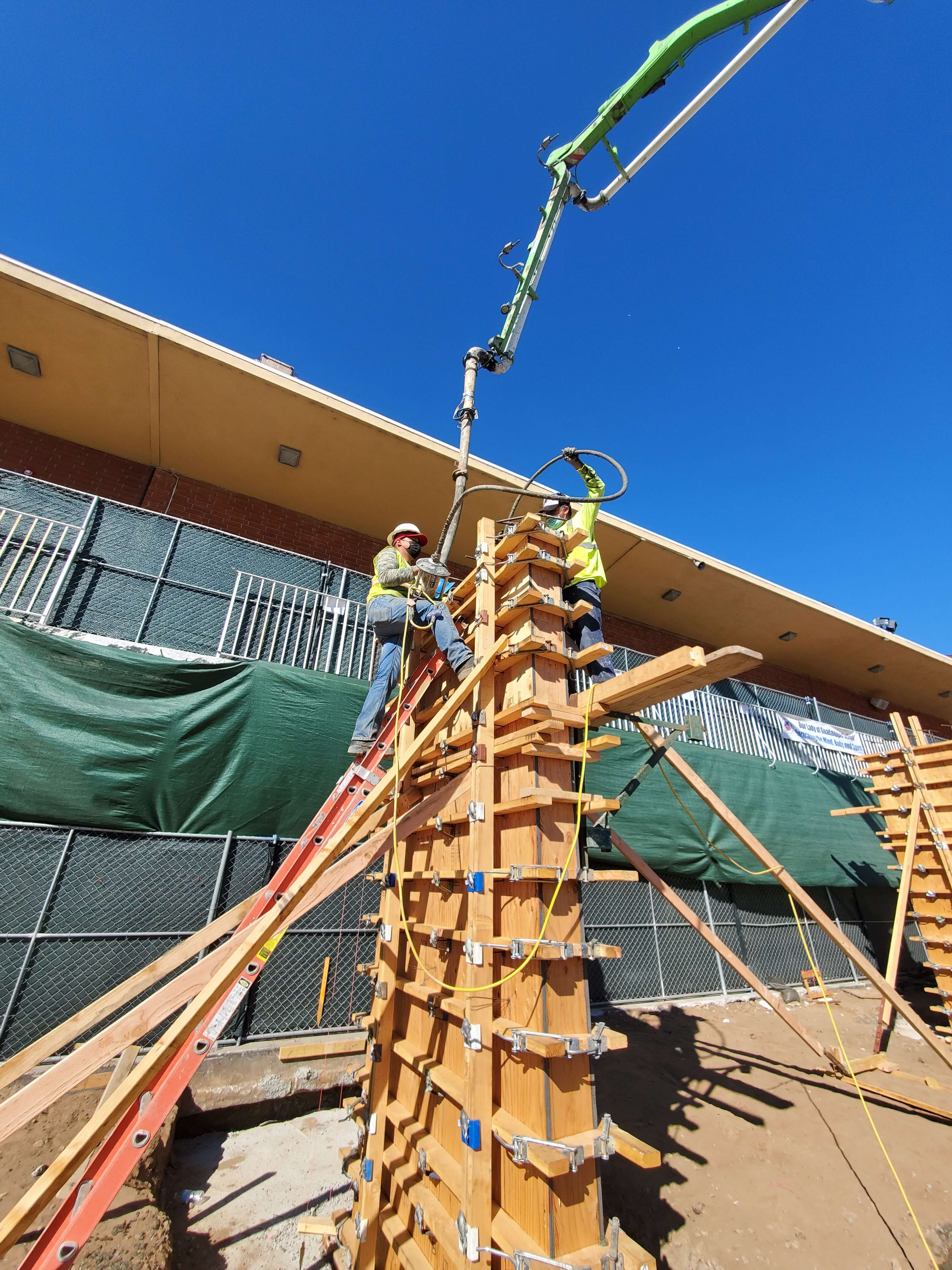 Two construction workers in safety gear operate a concrete pump hose to pour concrete into a wooden form structure on a construction site under a clear blue sky.