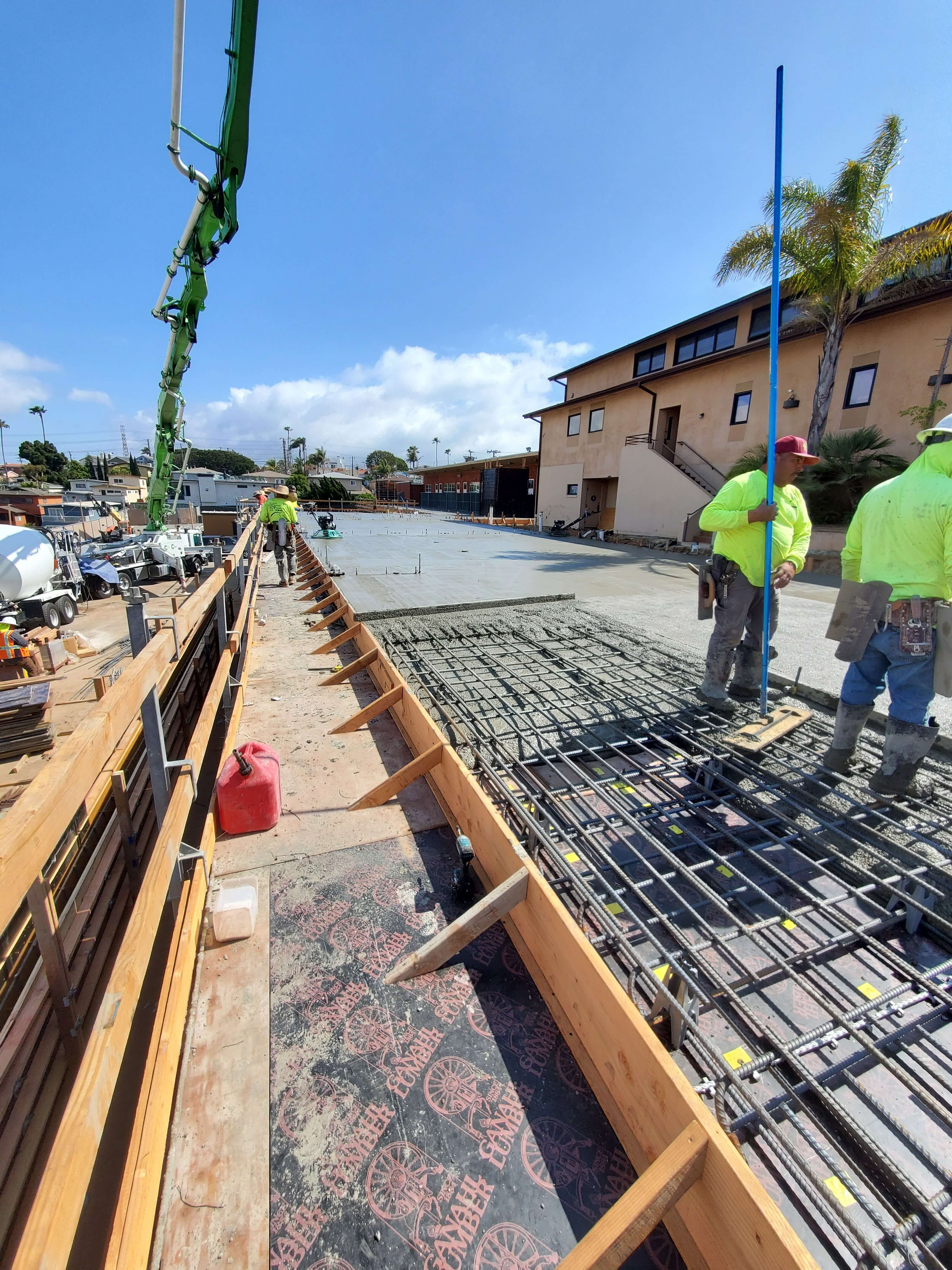 Construction workers in neon shirts pouring and leveling concrete over steel reinforcement near a residential building under a clear blue sky.