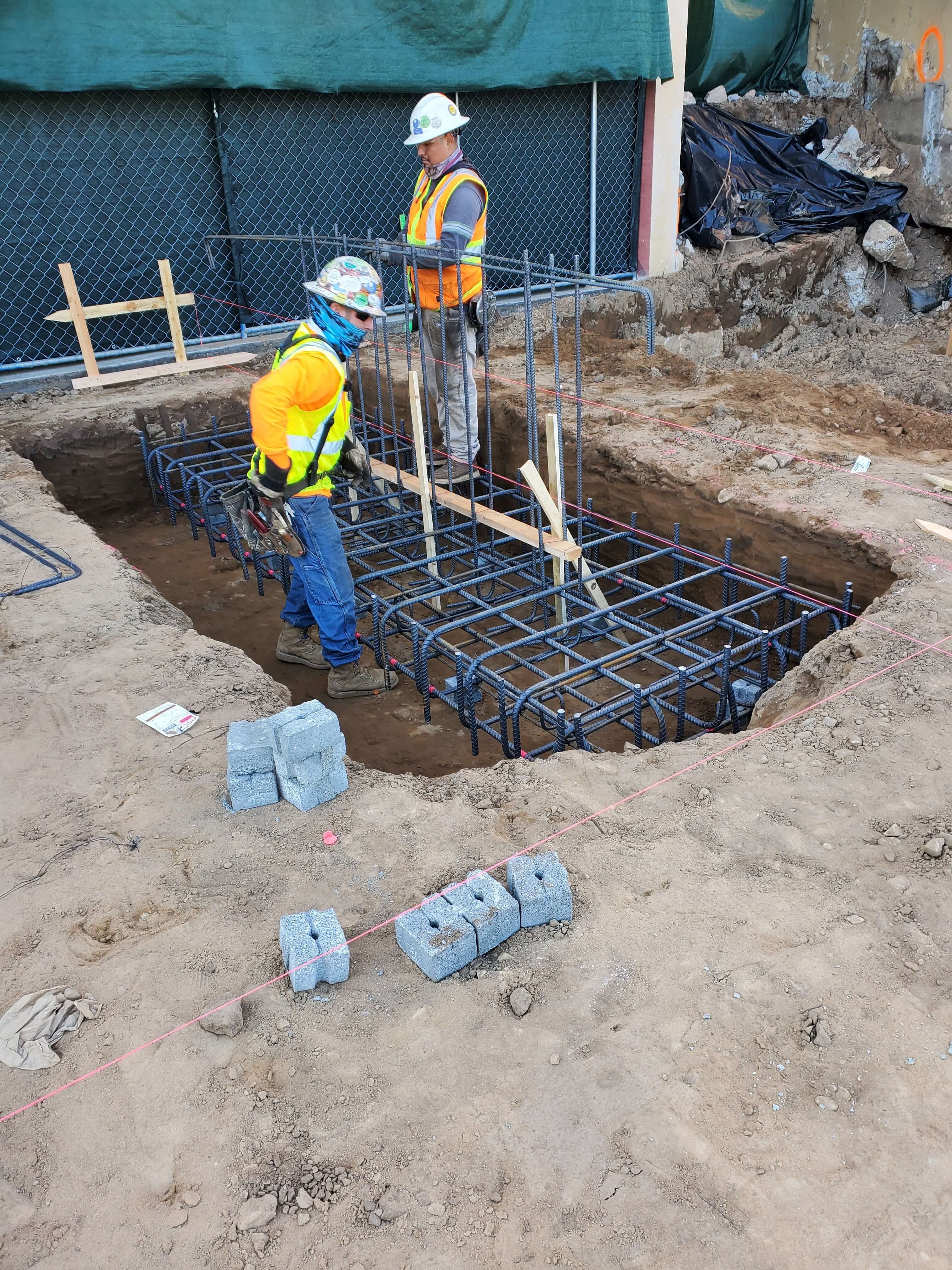 Two construction workers in safety gear working on steel rebar foundation inside a dug hole at a construction site.