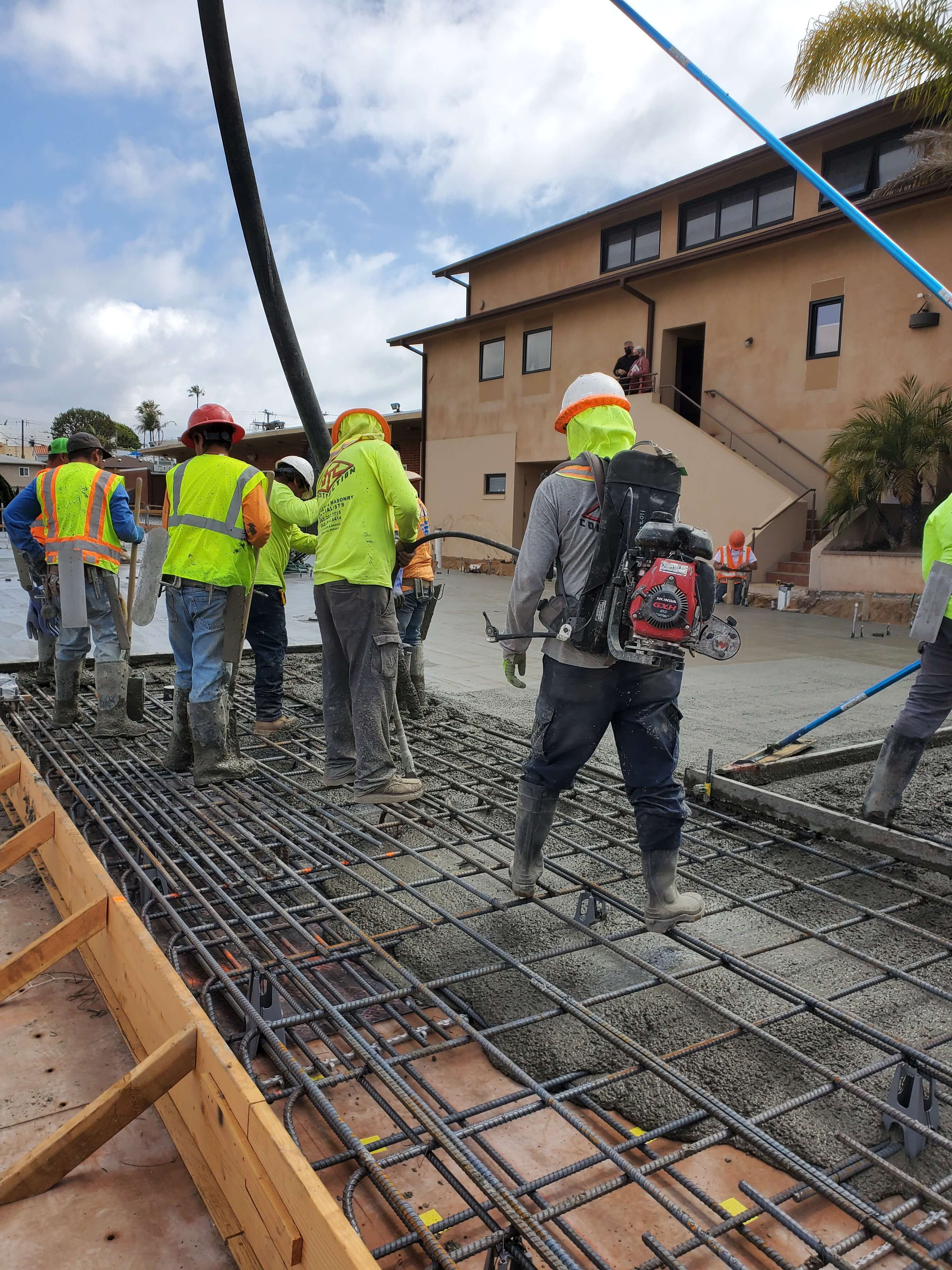 Construction workers in safety gear pouring and smoothing concrete over steel rebar at a building site.