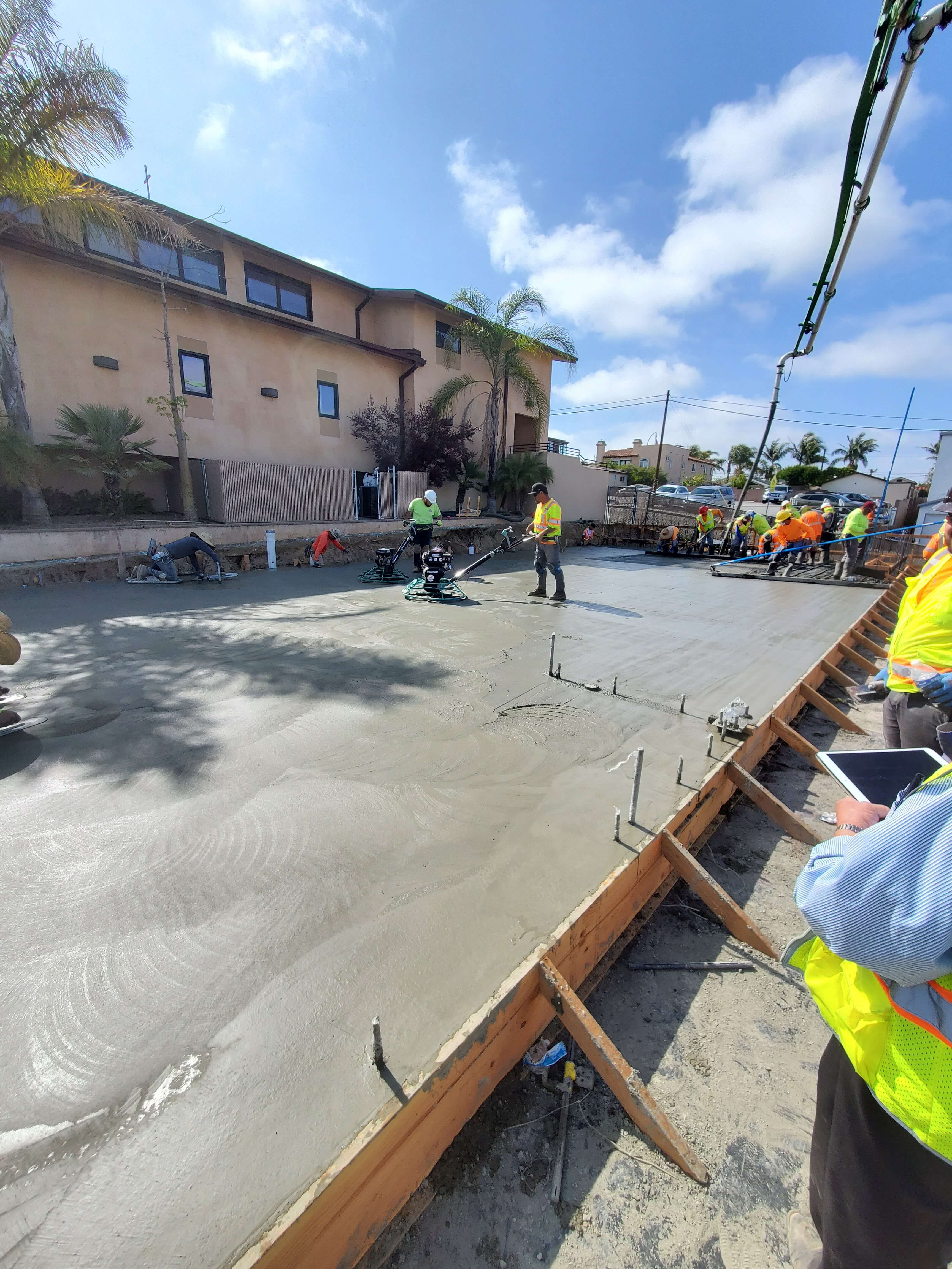 Construction workers in safety vests smoothing freshly poured concrete slab near residential buildings under a blue sky.