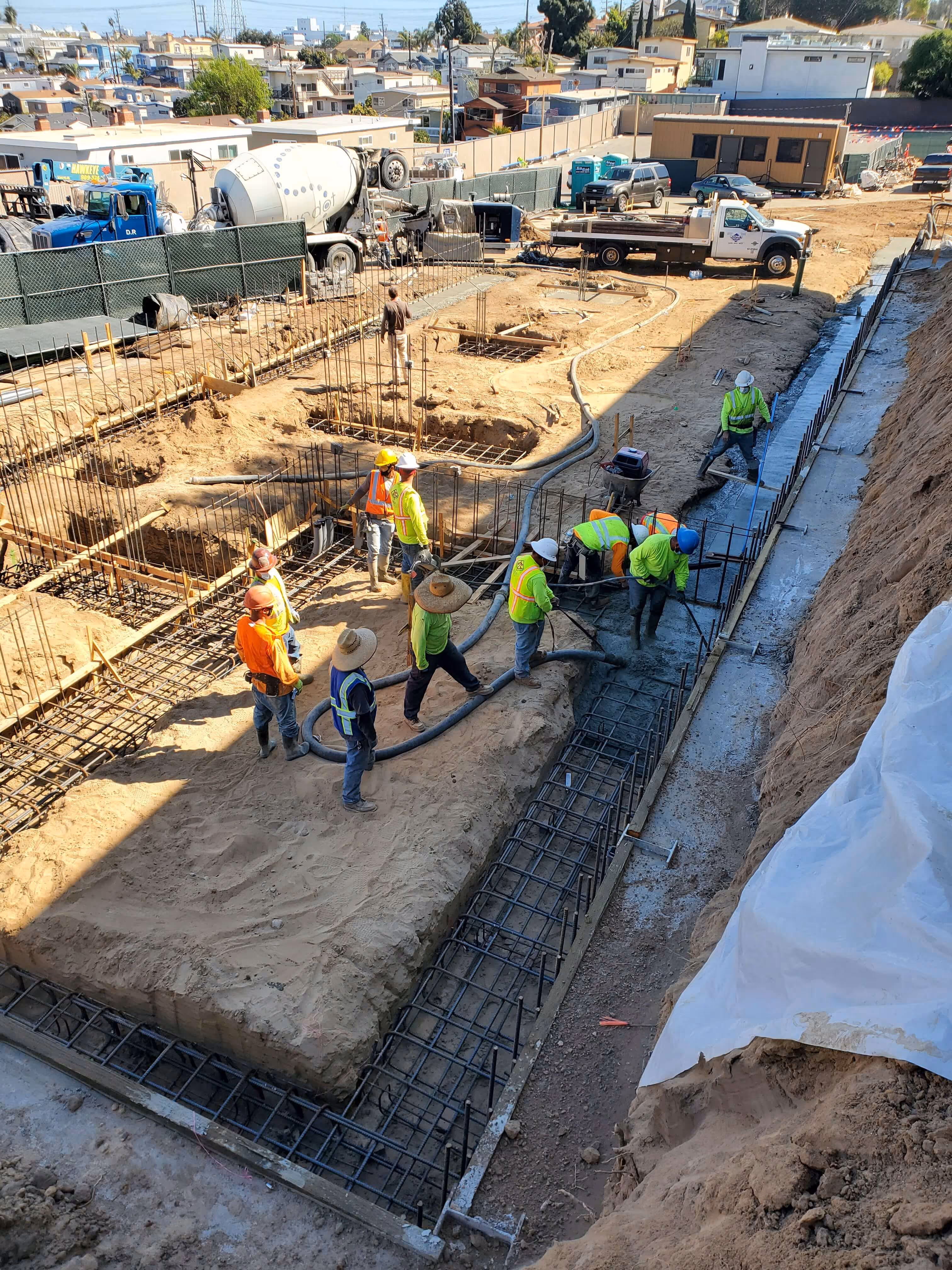 Two construction workers in safety vests and helmets pouring and spreading concrete over steel reinforcement mesh on a building site.