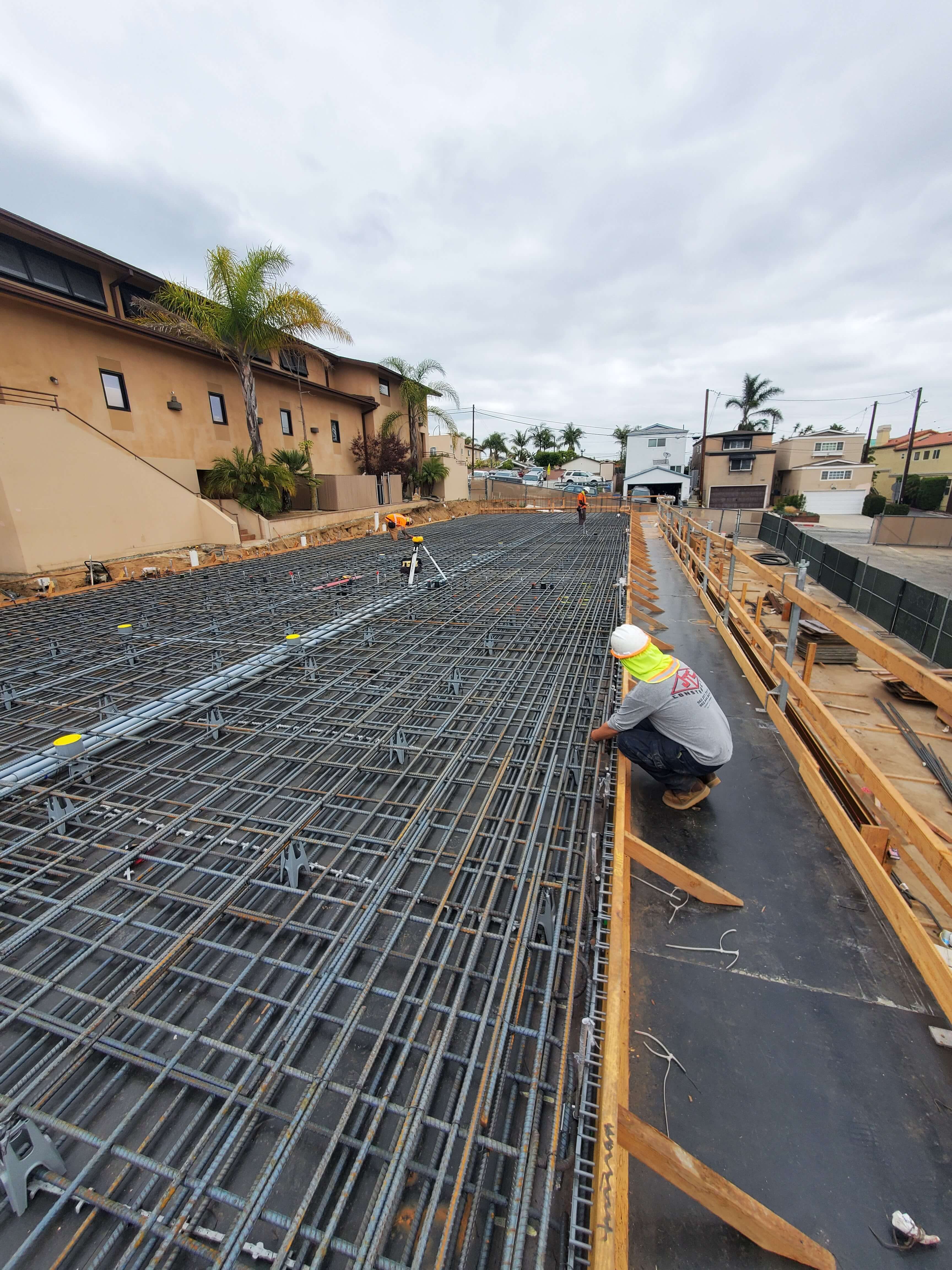 Construction site with workers installing metal rebar grid for concrete foundation near residential buildings under cloudy sky.