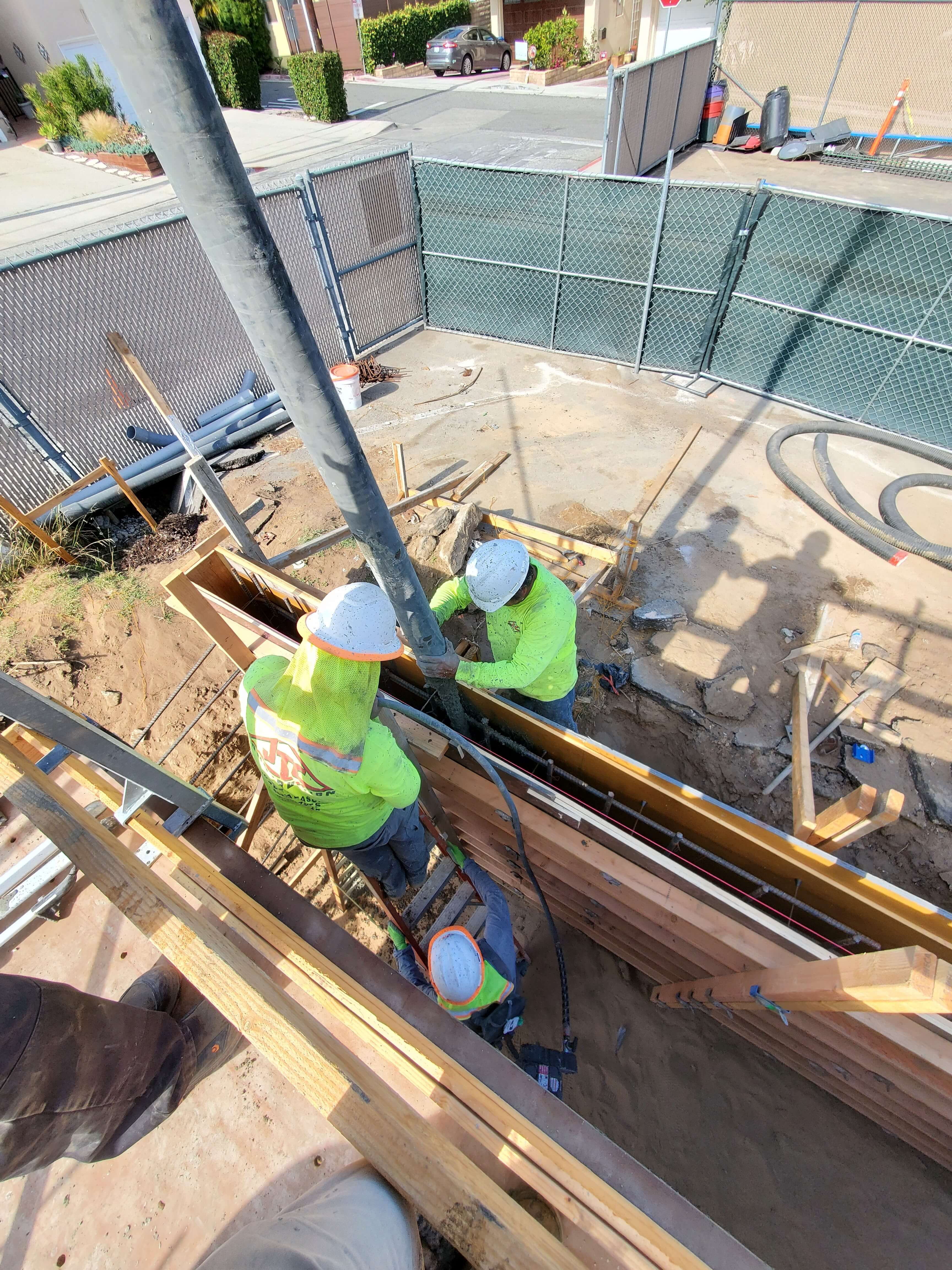 Three construction workers in safety helmets and neon clothing pouring concrete into a wooden formwork at a construction site.