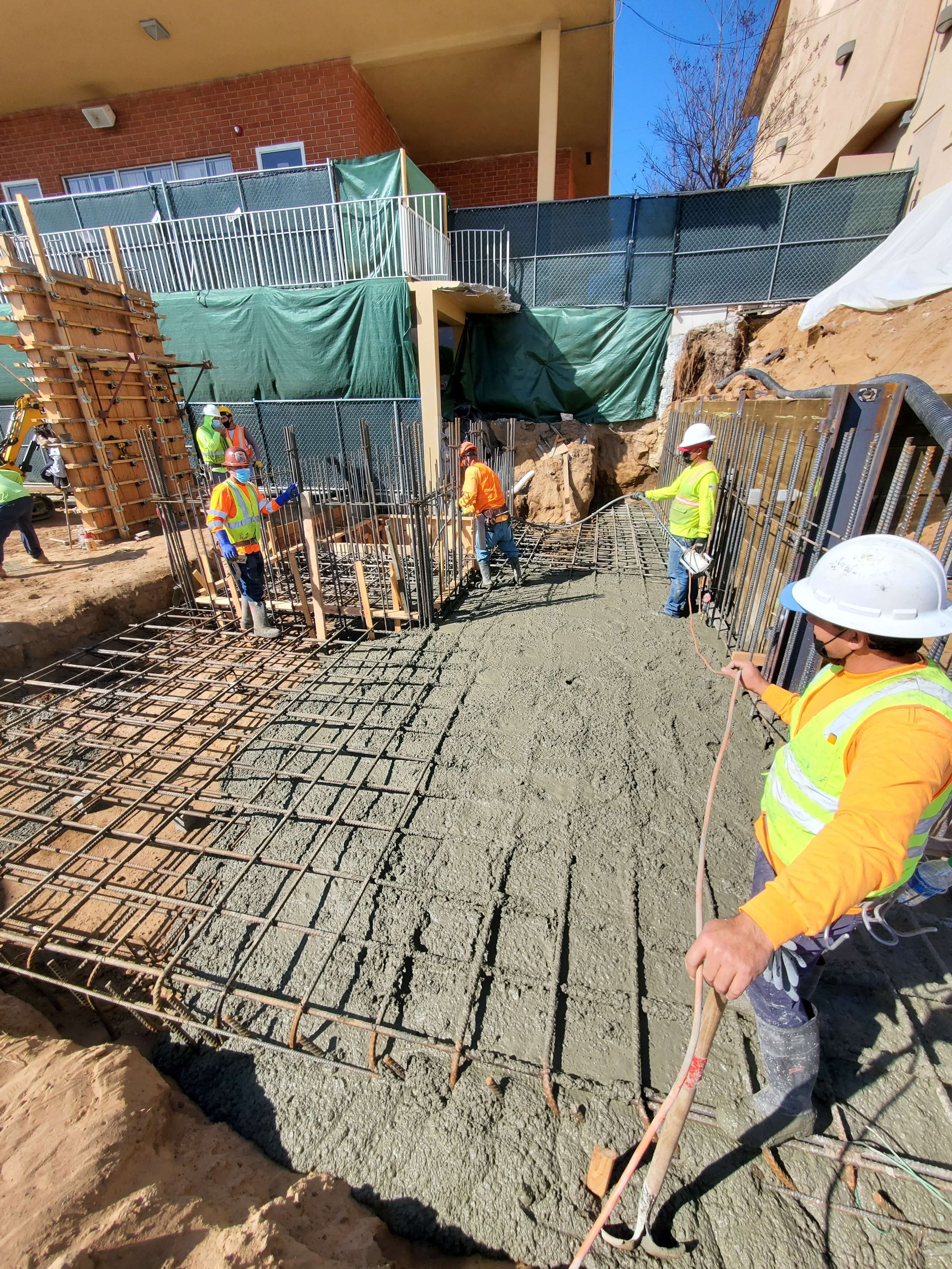 Construction workers in safety gear pouring and spreading concrete on a rebar framework at a building site.