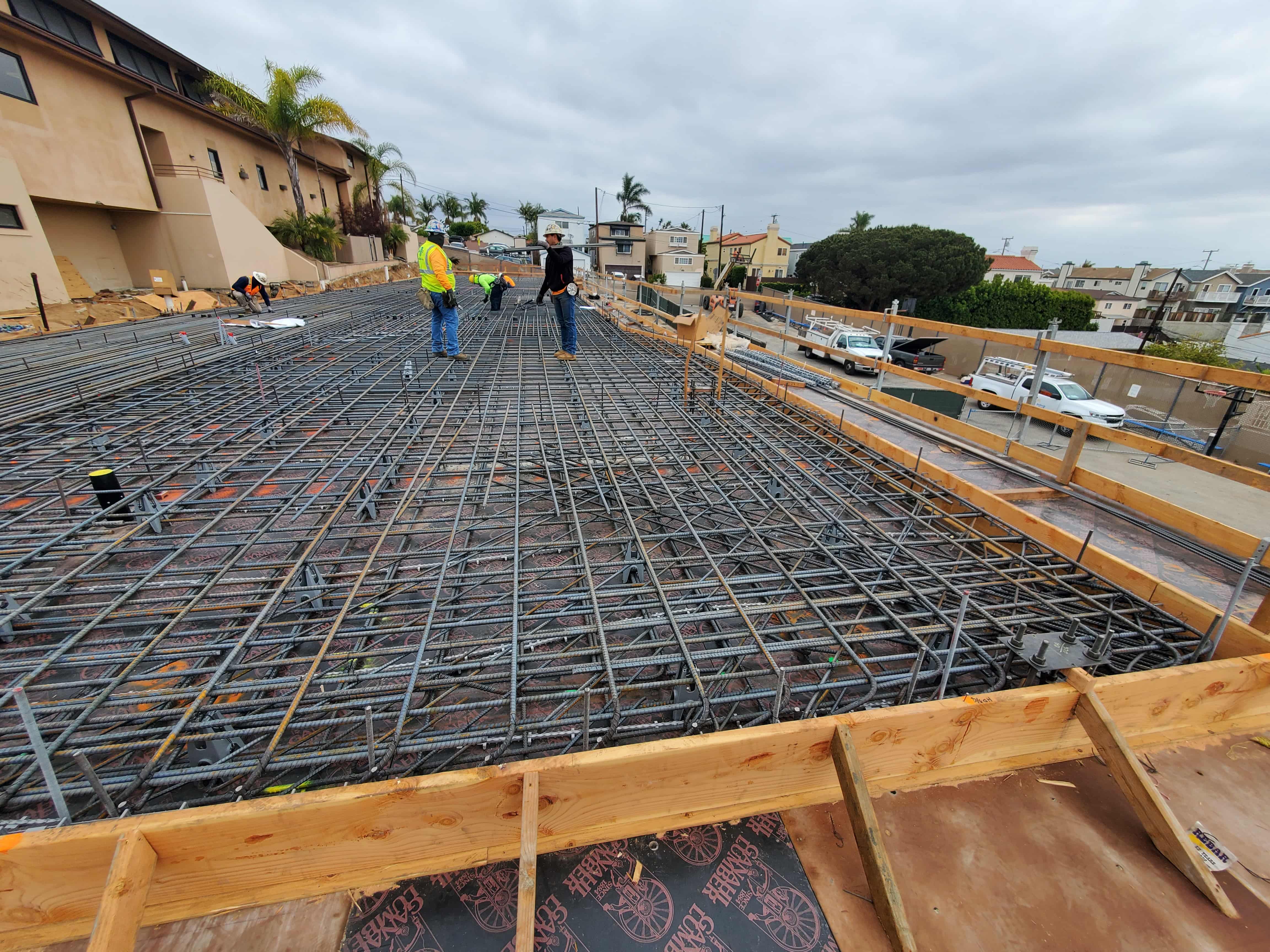 Construction workers wearing safety vests and helmets working on a large steel rebar grid for a concrete foundation under an overcast sky.