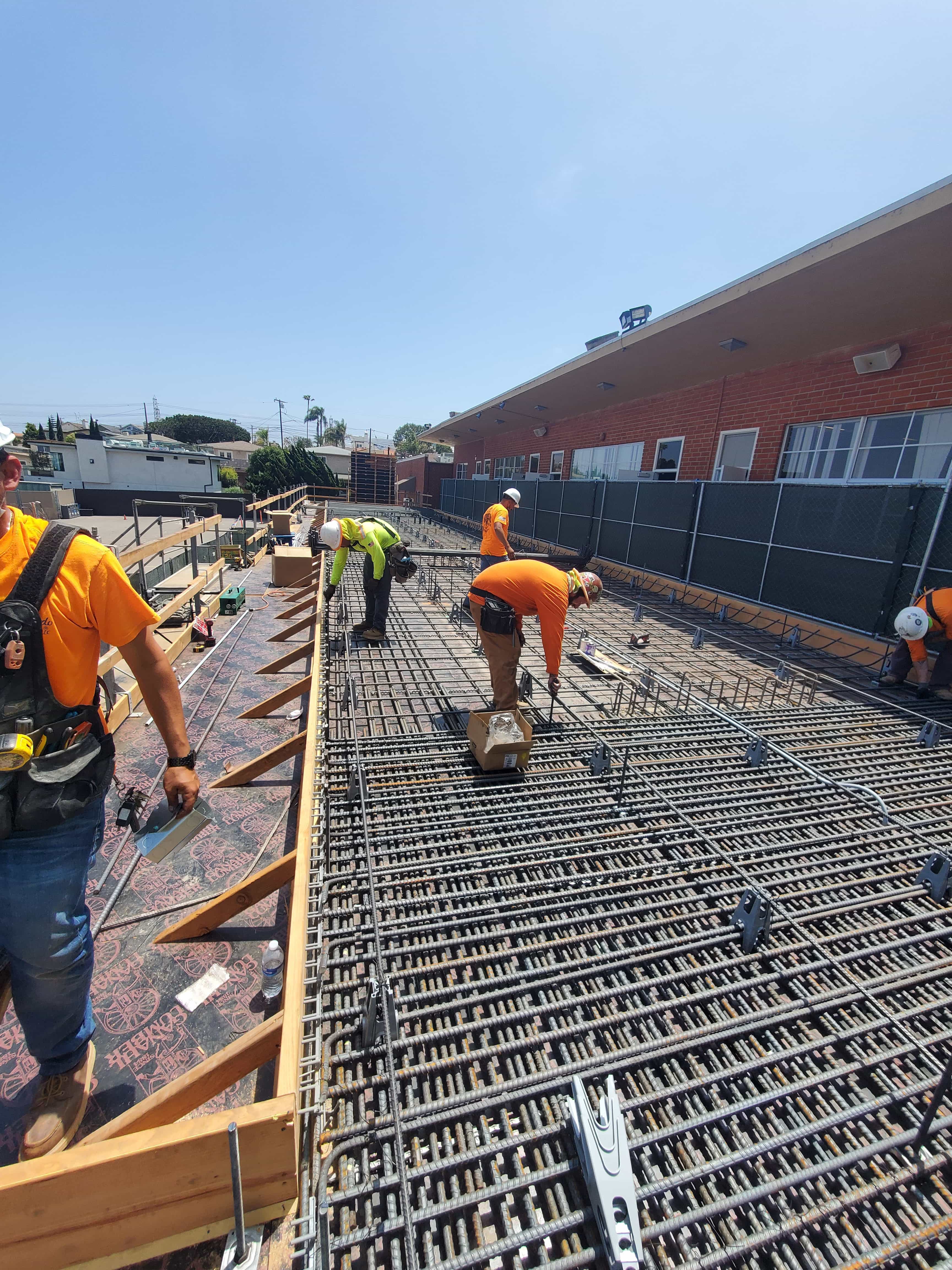 Construction workers wearing helmets and safety gear installing rebar on a building site under a clear blue sky.
