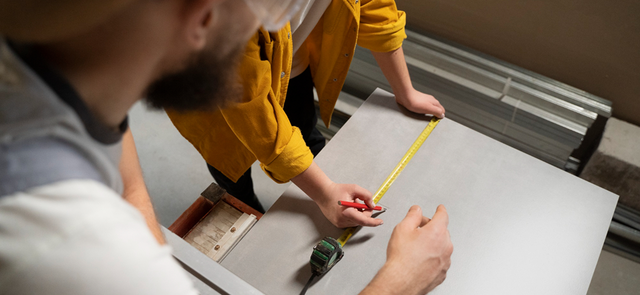 Two people measuring and marking on a flat board using a tape measure and pencil.