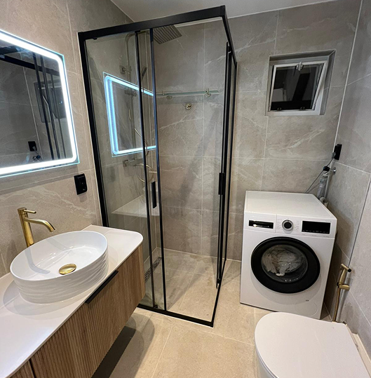 Modern bathroom with beige tiled walls and floor, a glass shower enclosure with black frame, a white vessel sink with gold faucet, a front-loading washing machine, and a white toilet.