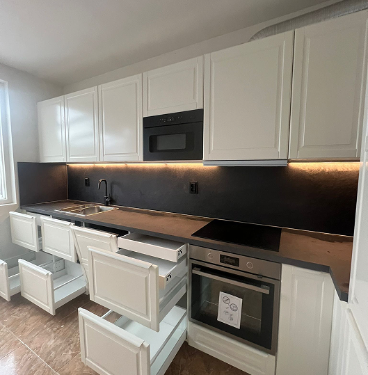 Empty modern room with light wooden floor, beige walls, a window with sheer and heavy curtains, and recessed ceiling lights.