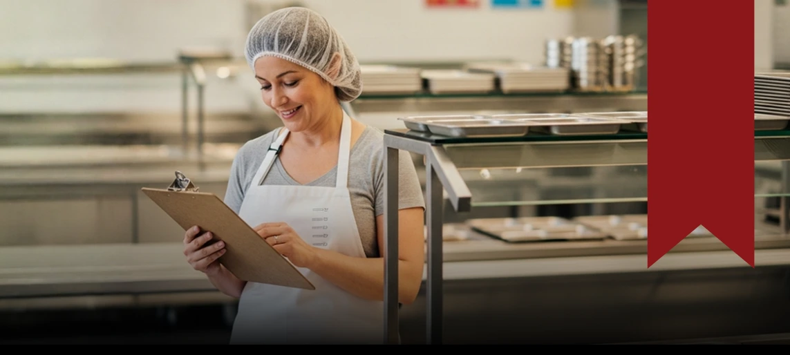 K-12 school kitchen staff member reviewing food safety checklist to support compliant meal preparation and inspection readiness.