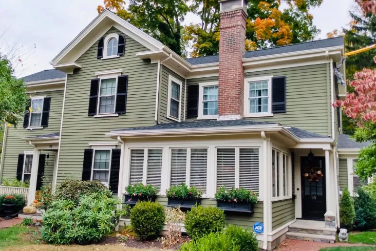Green two-story house with brick chimney and lush garden.
