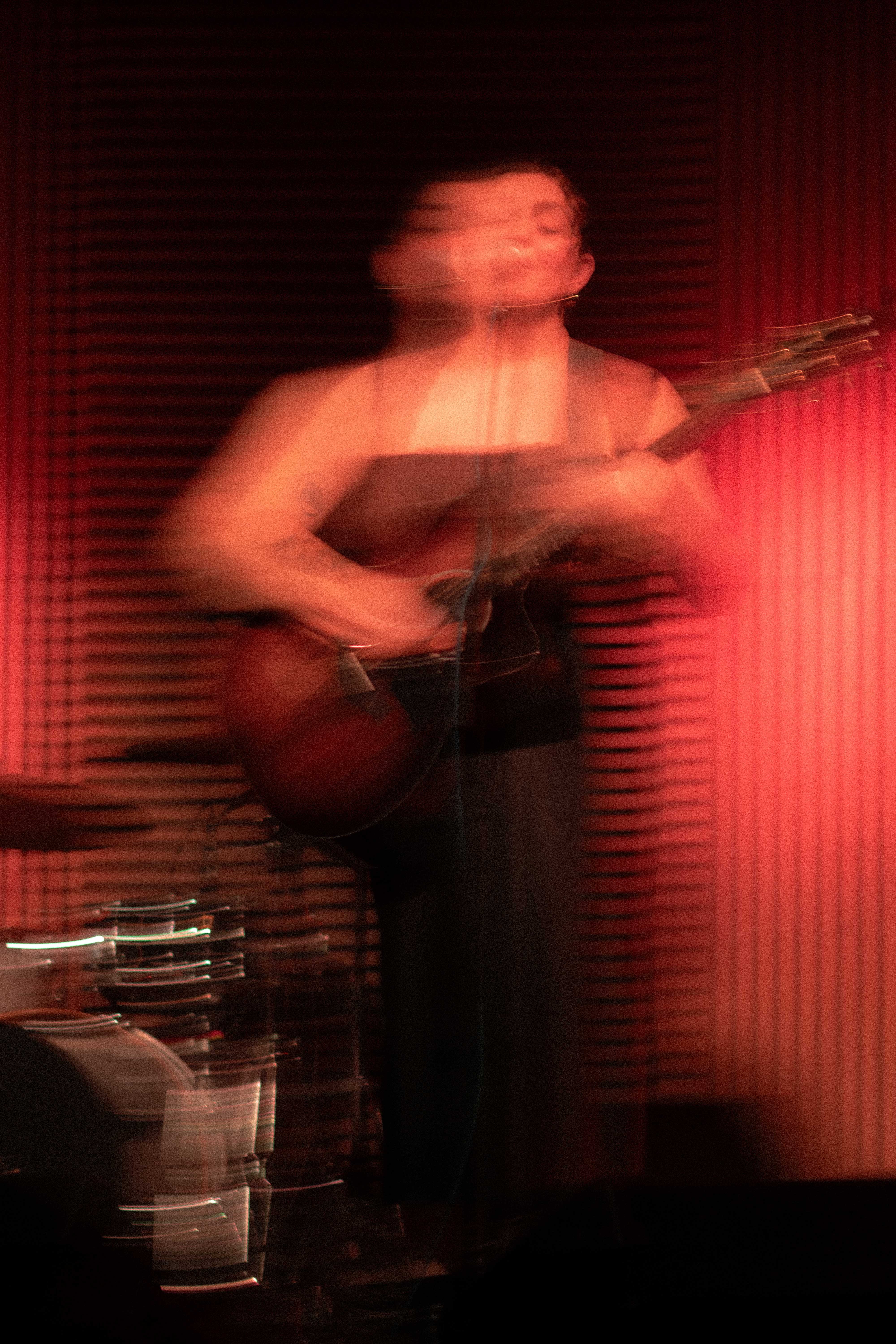 A young woman on a stage with red lighting behind her holding a guitar with motion blur affect.