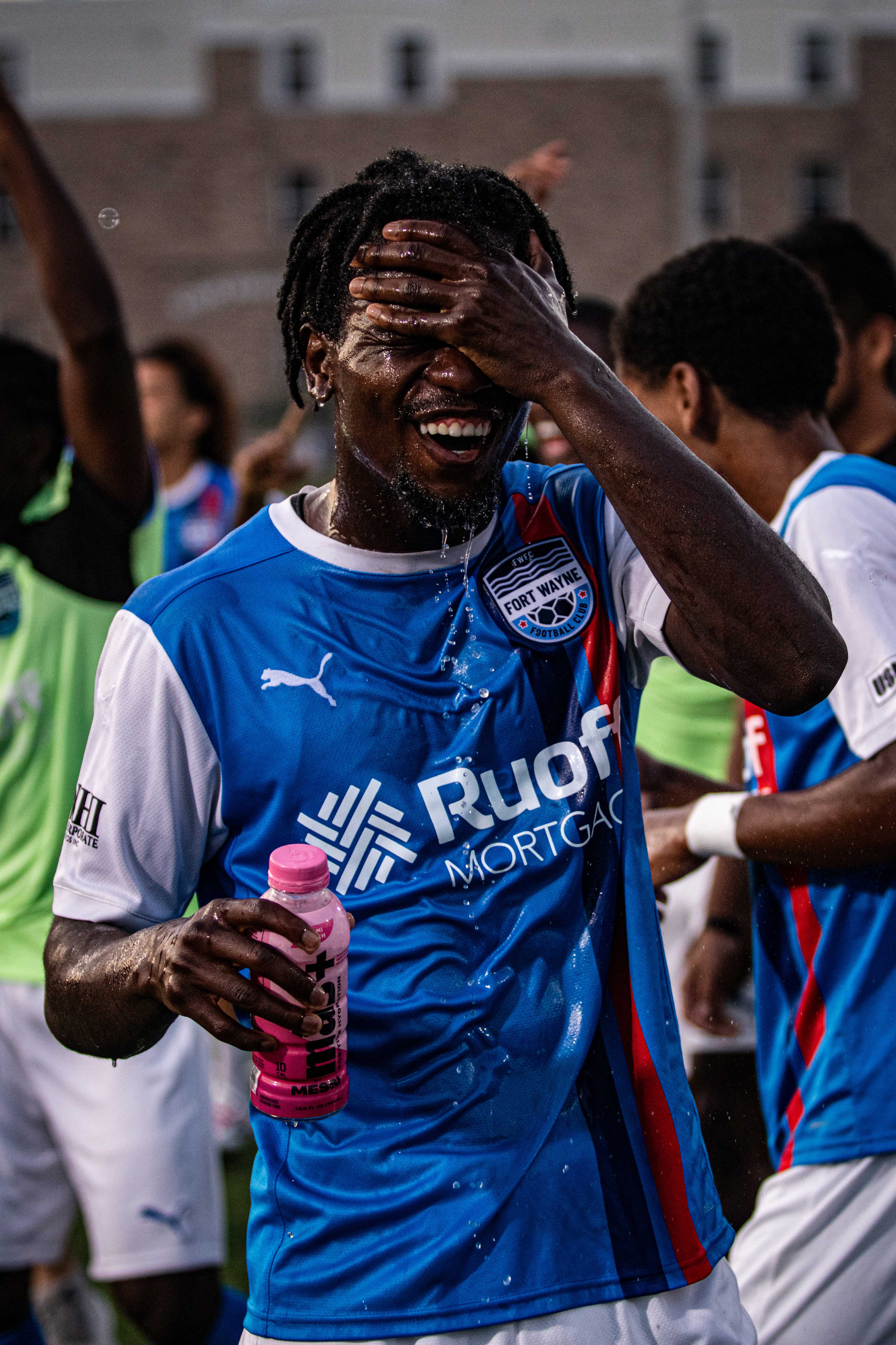 A young male athlete pour water on his face and smiling in a blue jersey.