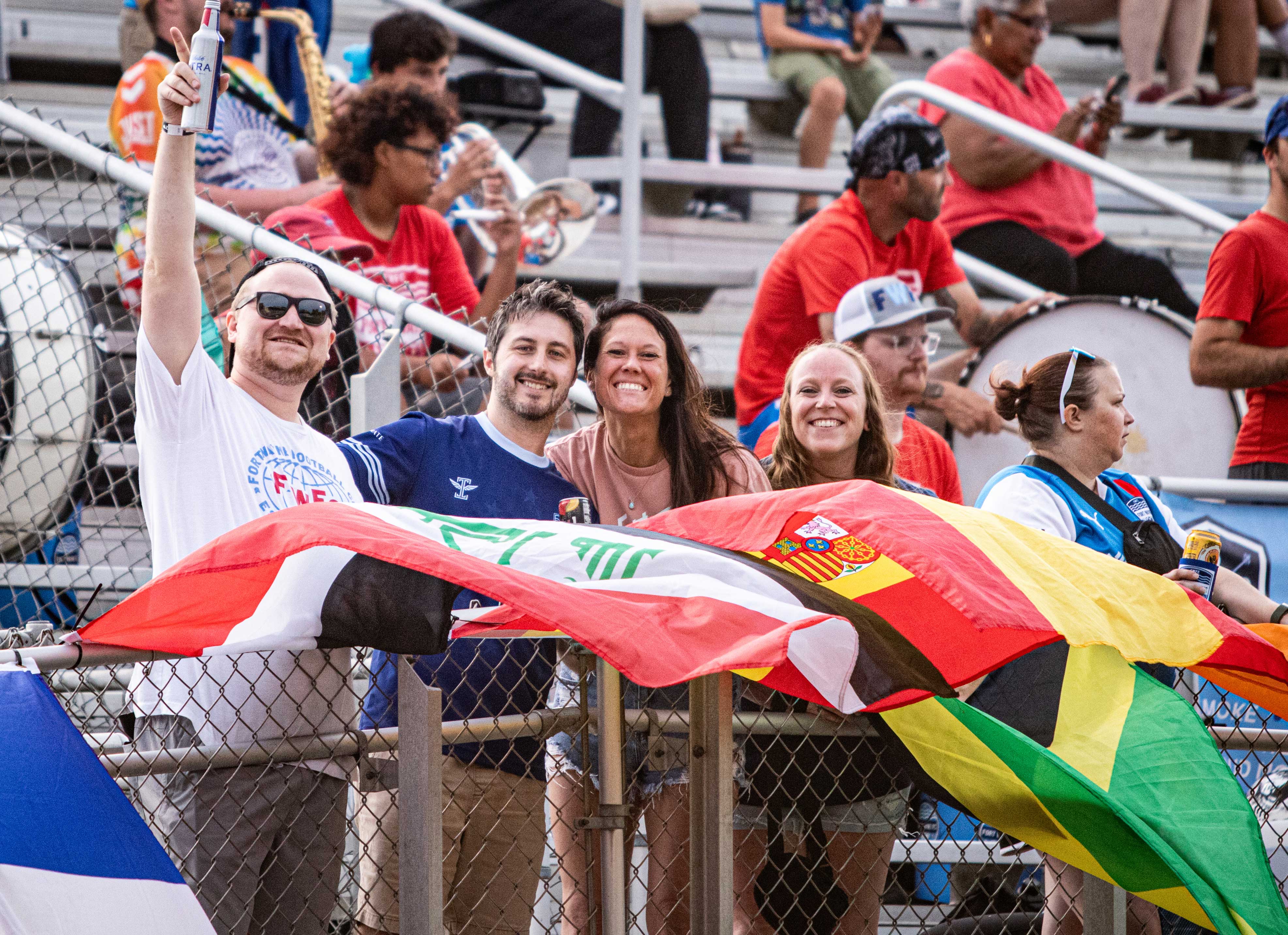 4 soccer fans smiling at the camera at the front of the stands with flags waving in the wind.