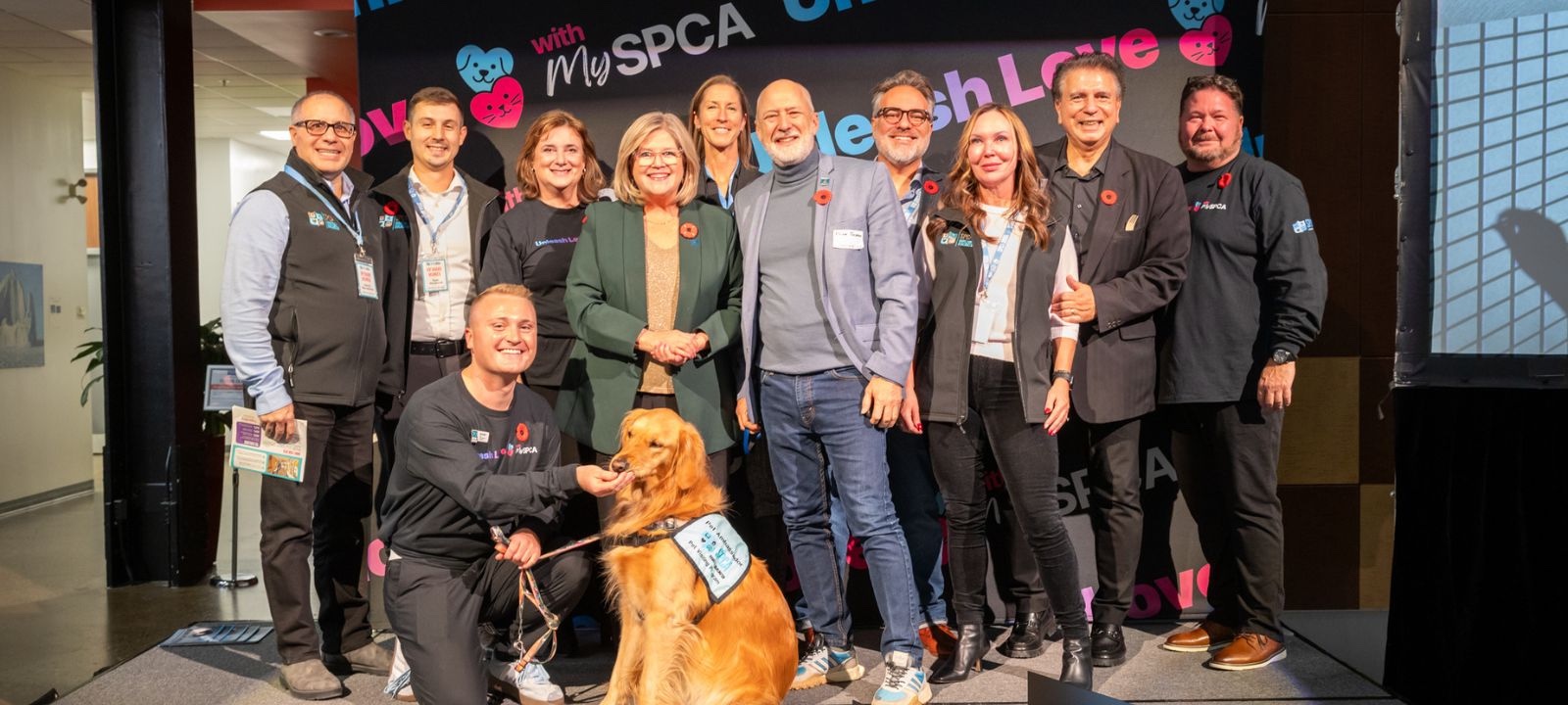 Group photo of eleven people smiling on a stage with a golden retriever wearing a service dog vest, and a MySPCA backdrop behind them.