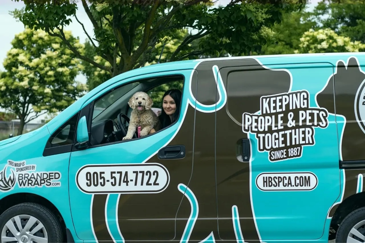 Blue and brown van with animal outlines and text 'Keeping People & Pets Together Since 1887,' a woman and a small dog sitting inside the driver's window.