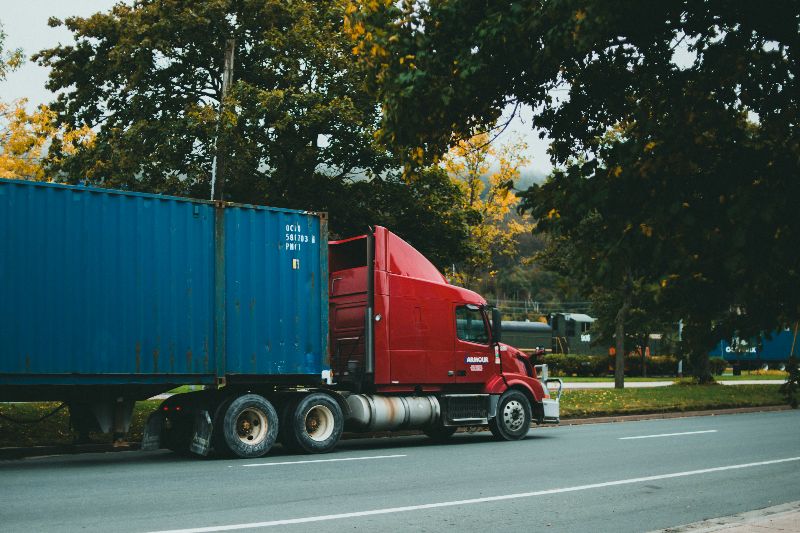 Red semi-truck with blue shipping container parked roadside under trees with autumn leaves, transporting freight on a city street.