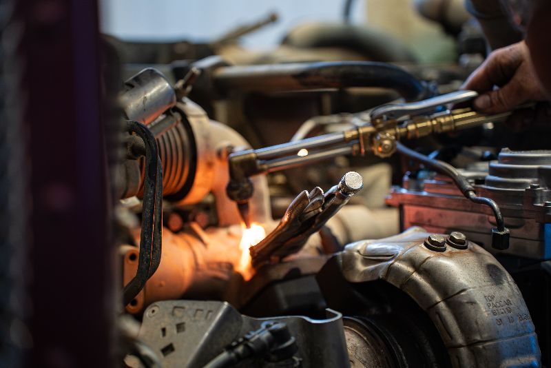 Mechanic using a torch to heat a metal component inside a diesel engine bay, with glowing flame and surrounding engine parts visible