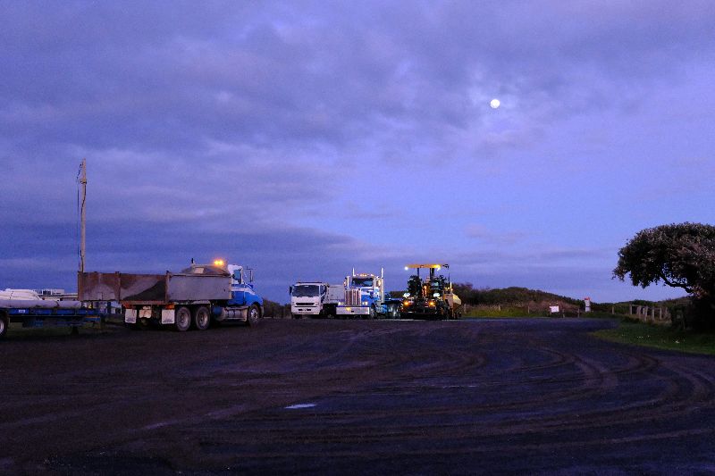 Parked construction trucks and heavy vehicles line a quiet roadside at dusk beneath a cloudy sky with a visible moon.